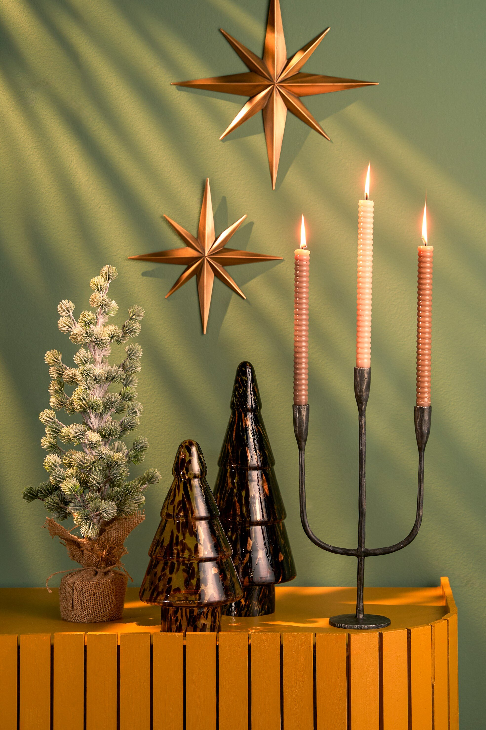 Scène festive avec des étoiles, des bougies et des arbres décoratifs sur une étagère jaune contre un mur vert.