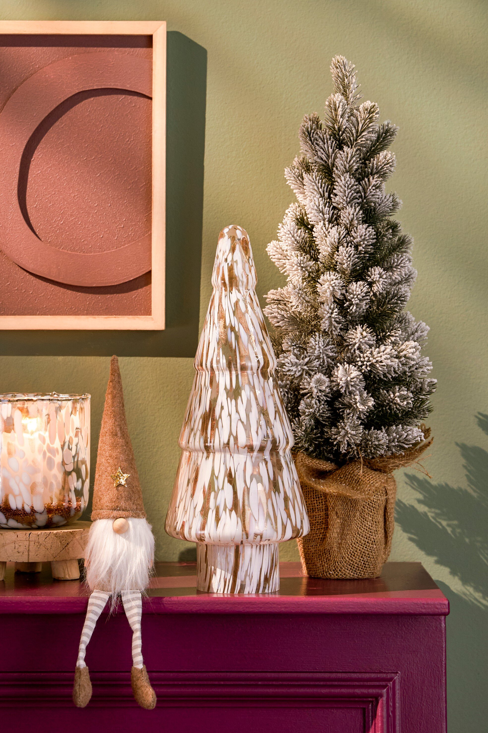 Scène festive : Sapin de Noël décoré, arbre en verre, gnome et bougie sur une surface violette. Fond vert.