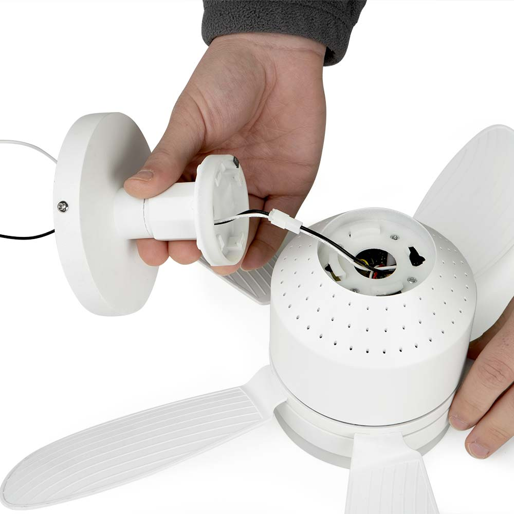 Una persona montando un ventilador de techo blanco. Las manos conectan cables. Se ven las aspas y la carcasa del motor. Manga gris.