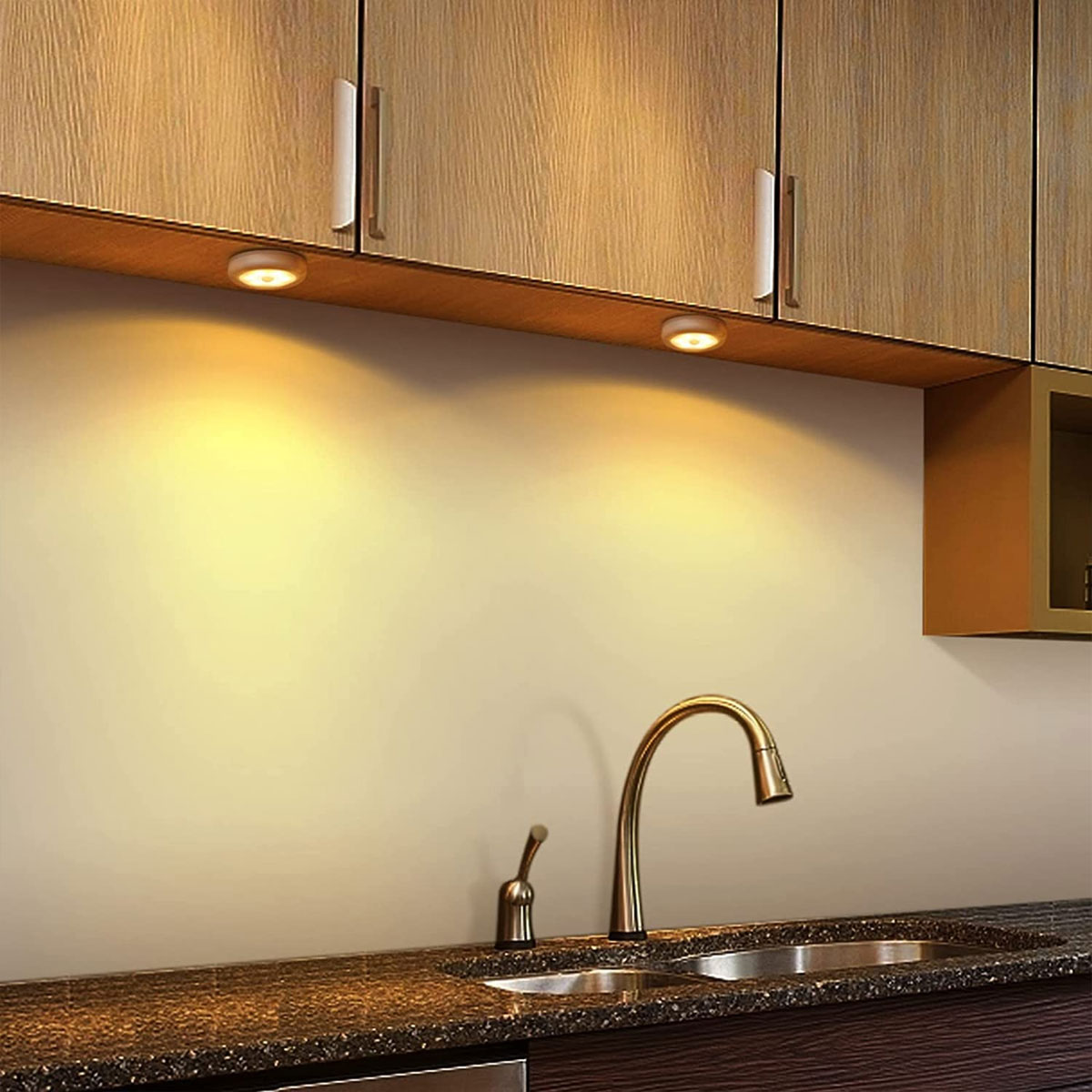 Eine Küchenszene mit Holzschränken, einem Waschbecken und einem Wasserhahn. Zwei Lichter beleuchten die Wand. Die Arbeitsplatte ist dunkel.