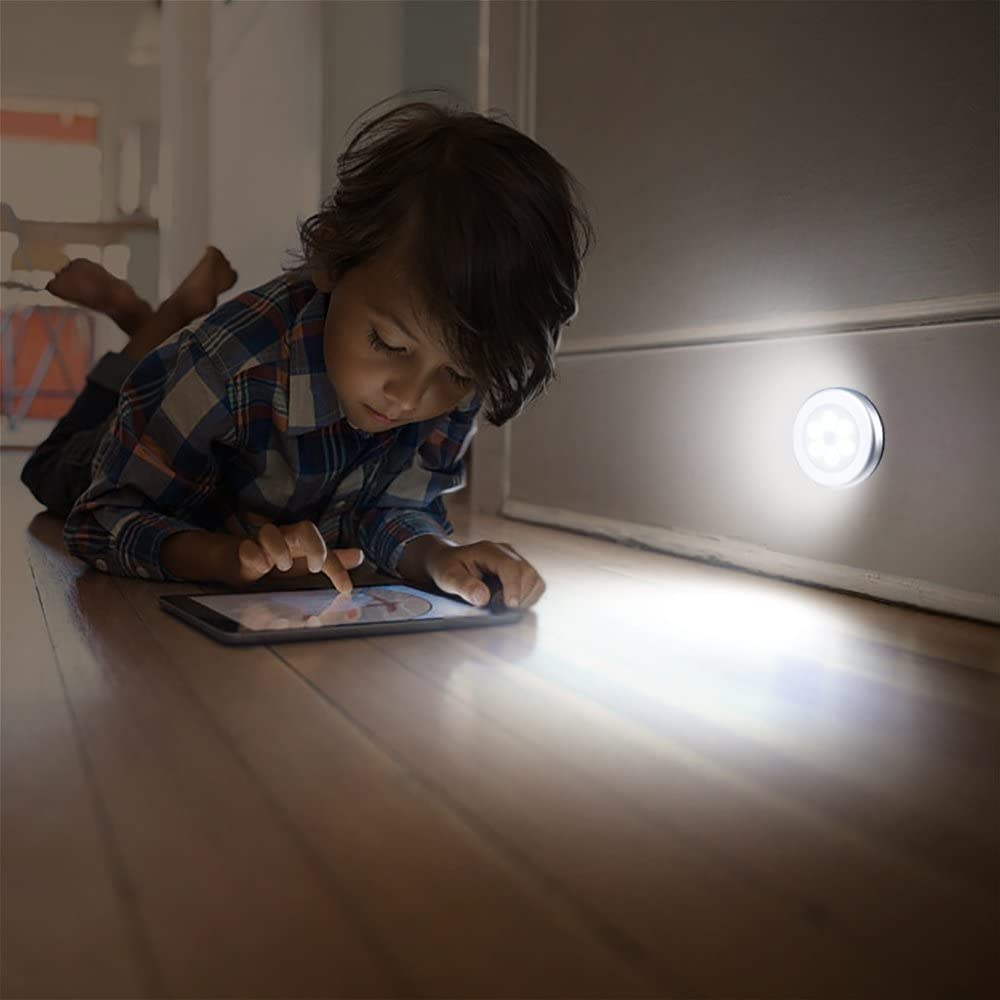 Ein Kind liegt auf einem Holzboden und benutzt ein Tablet. Ein helles Licht beleuchtet die Szene, und eine kleine Lampe ist an der Wand.