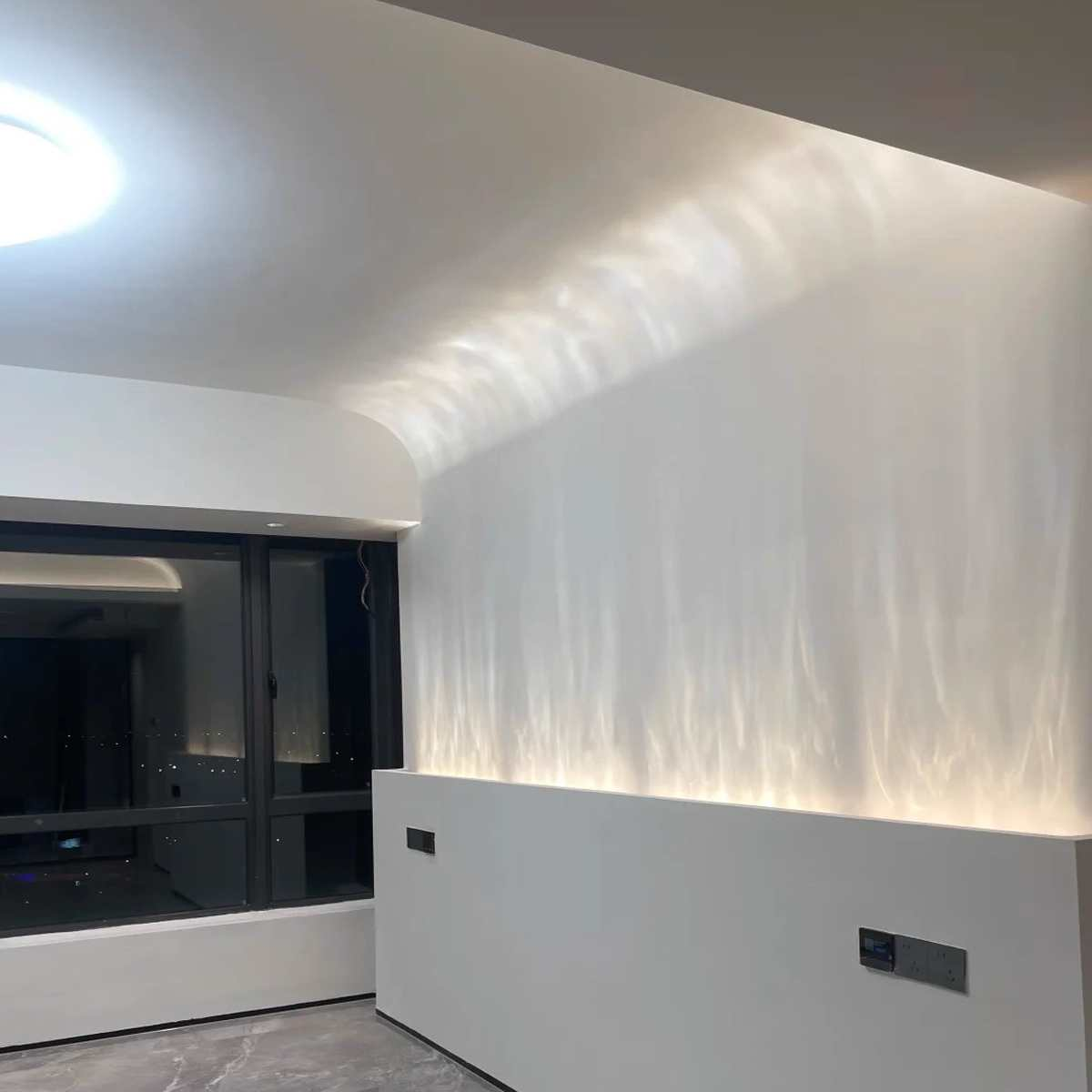 Weiße Wand mit Lichtreflexen. Ein Fenster links. Steckdosen sind an der Wand zu sehen.