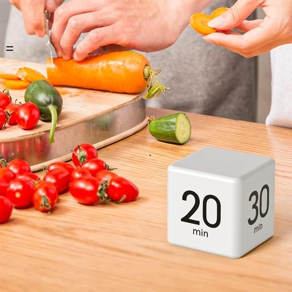 Eine Person schneidet eine Karotte auf einem hölzernen Schneidebrett. Cherrytomaten, eine Gurke und ein Timer-Würfel sind auch auf dem Tisch.