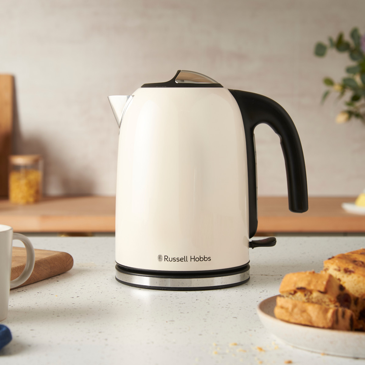 Ein cremefarbener Russell Hobbs Wasserkocher mit schwarzem Griff steht auf einer weißen Arbeitsplatte. Geschnittenes Brot ist im Vordergrund.