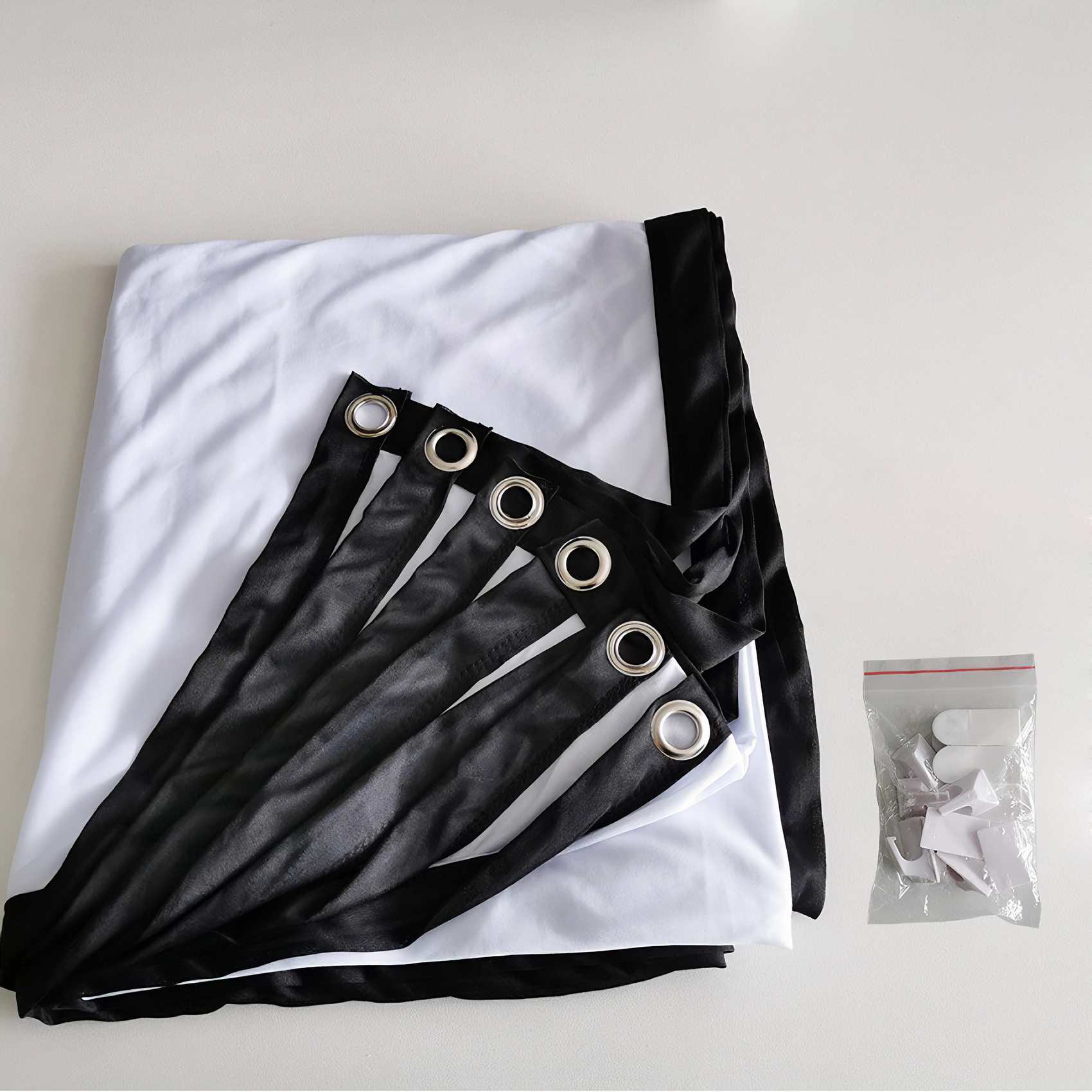 Eine weiße Projektorleinwand mit schwarzen Rändern und Metallösen sowie eine kleine Tüte mit weißen Clips.