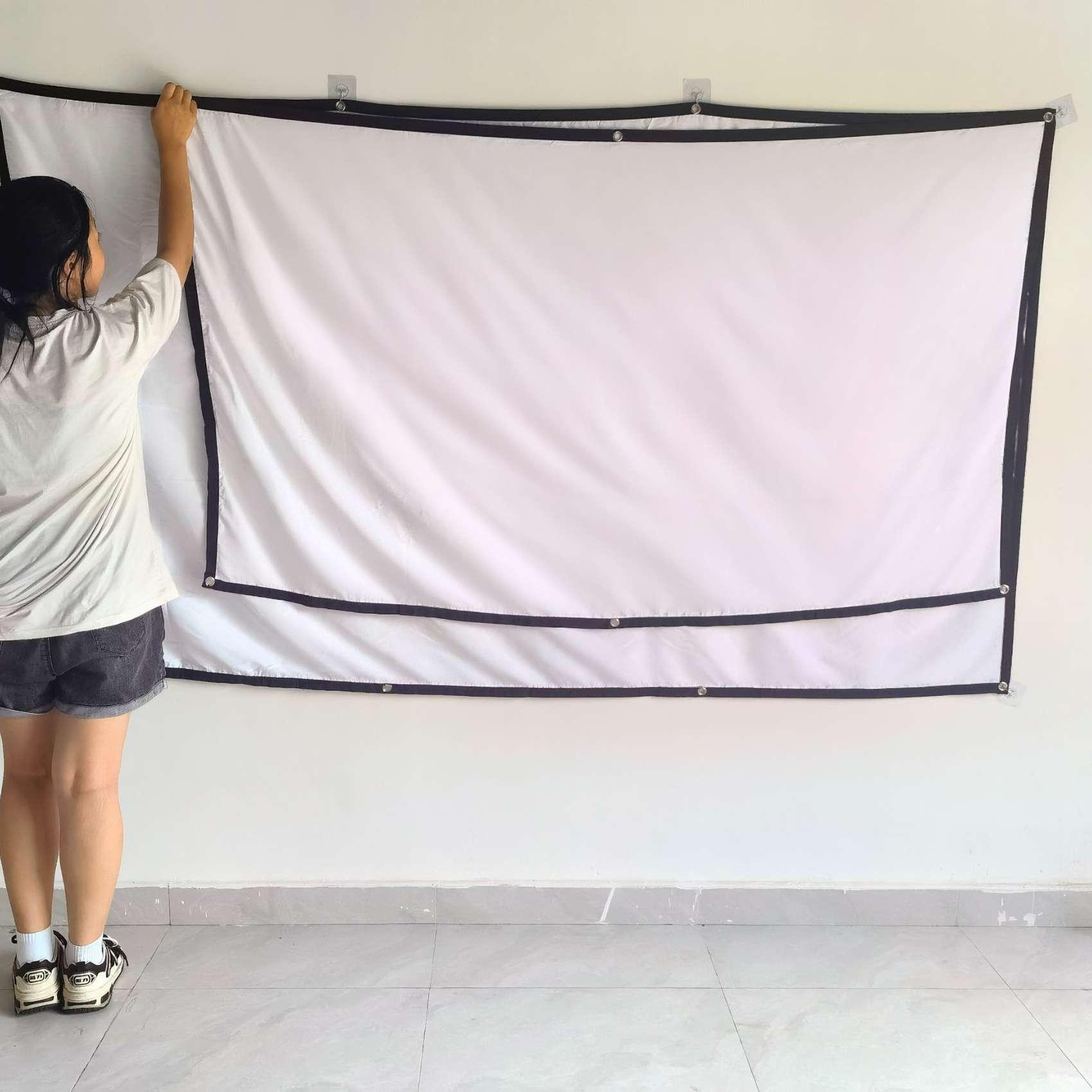 Eine Person hängt eine weiße Projektorleinwand an eine weiße Wand. Der Bildschirm hat schwarze Ränder. Die Person trägt Shorts.