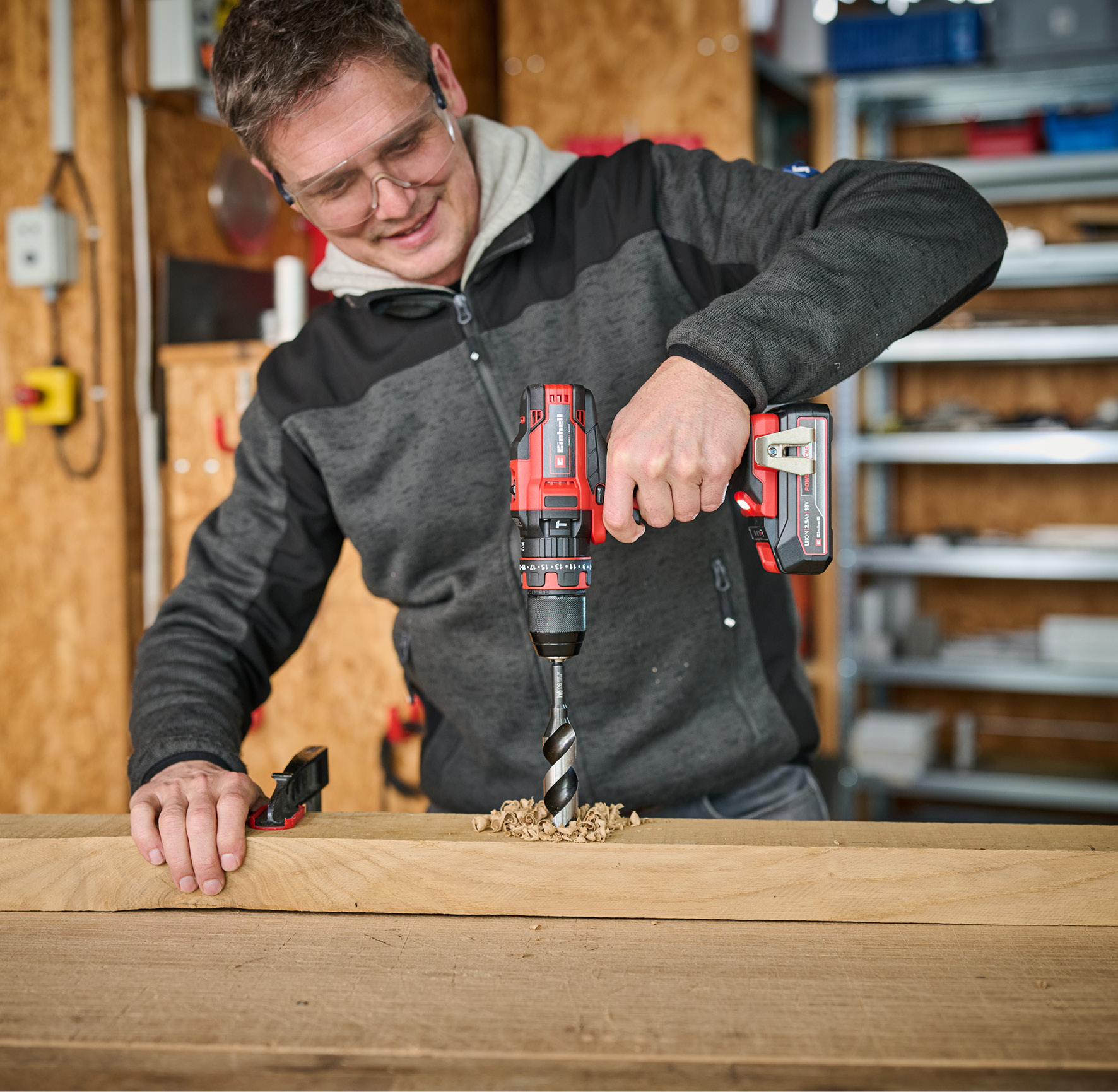 Mężczyzna wierci w drewnie wiertarką. Ma okulary ochronne. Widoczne trociny.