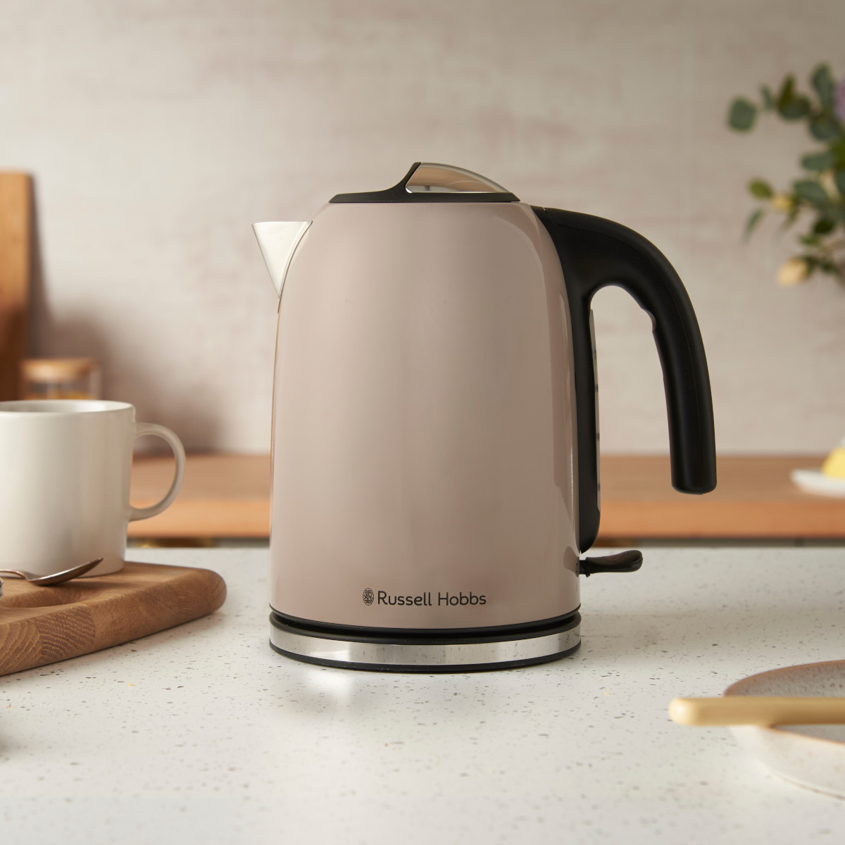 Ein beigefarbener Russell Hobbs Wasserkocher steht auf einer Arbeitsplatte. Links befinden sich eine weiße Tasse, ein Löffel und ein Holzbrett.