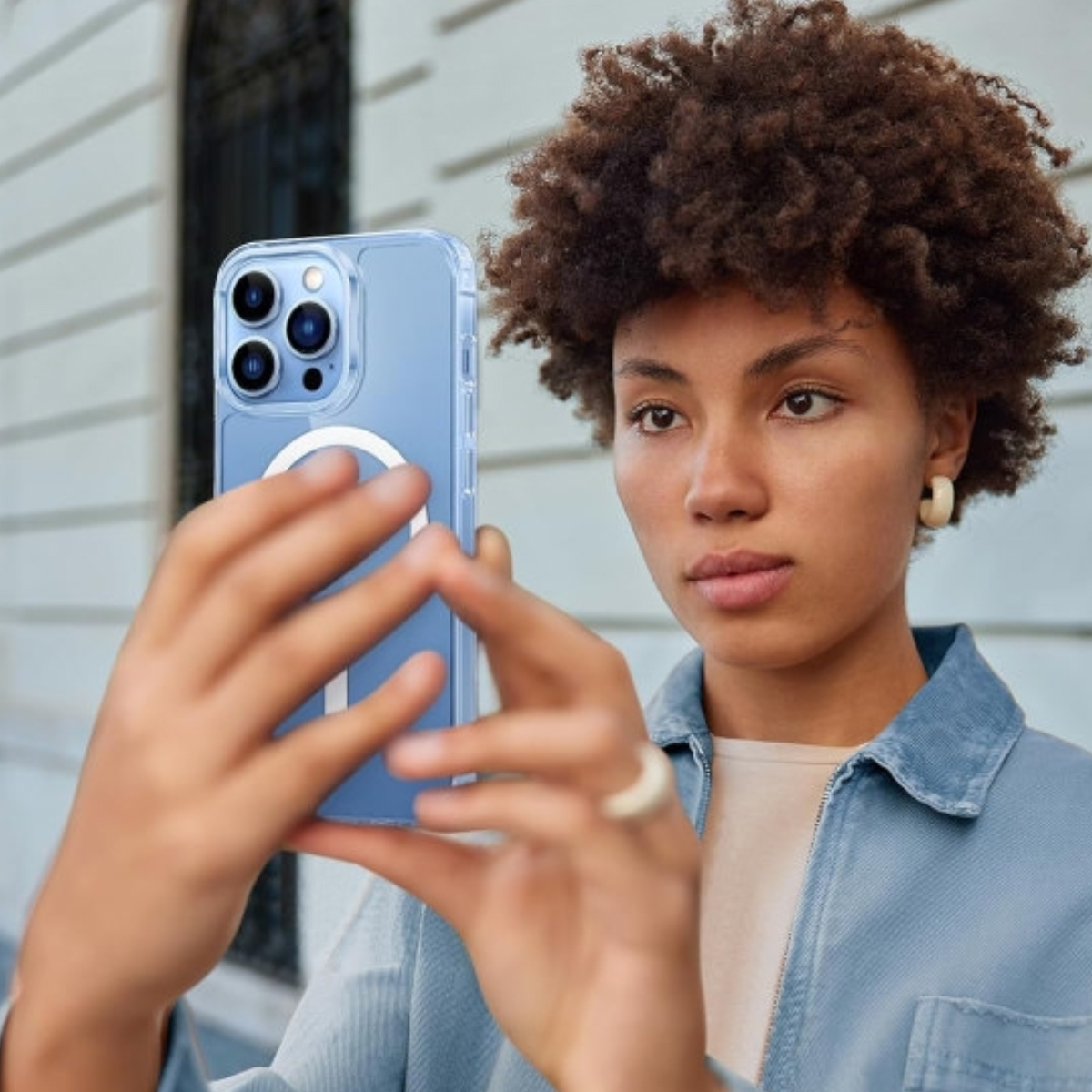 Eine Frau hält ein blaues iPhone. Sie trägt eine Jeansjacke und hat lockiges Haar. Der Hintergrund ist ein weißes Gebäude.