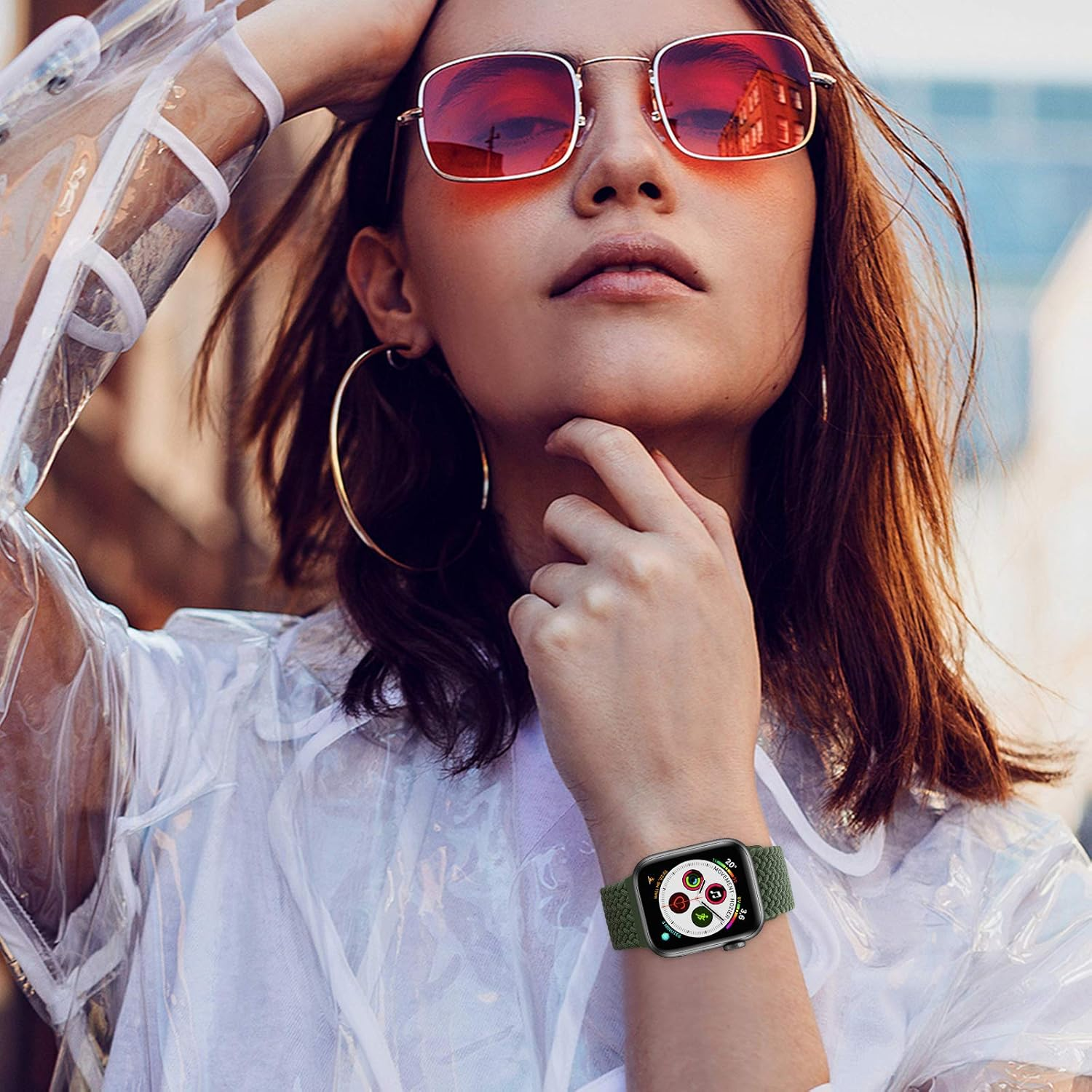 Eine Frau mit Sonnenbrille und Smartwatch. Sie trägt einen durchsichtigen Regenmantel und goldene Creolen. Gebäude spiegeln sich in den Gläsern.