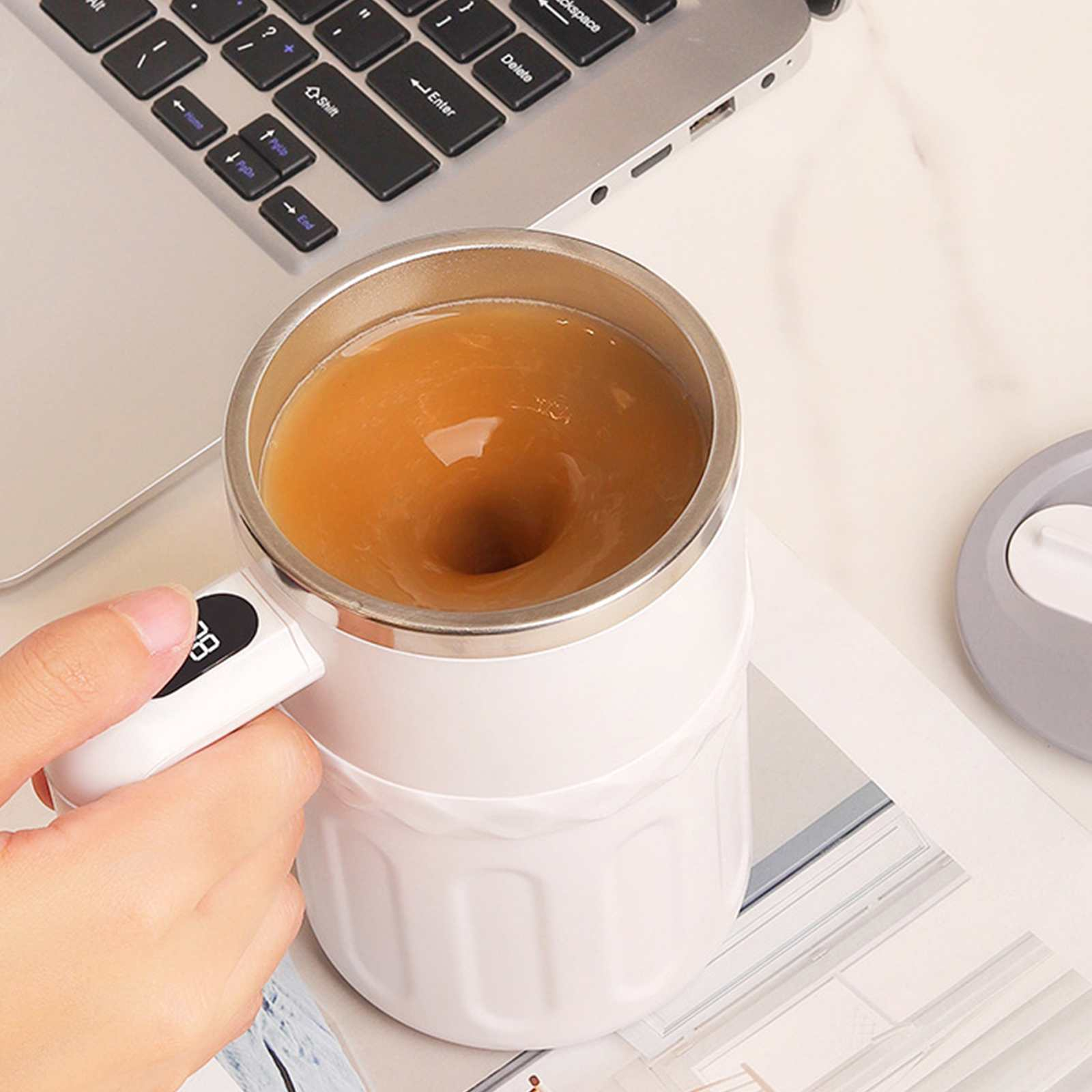 Ein weißer, selbstverrührender Becher mit Flüssigkeit steht auf einer weißen Oberfläche neben einem Laptop.
