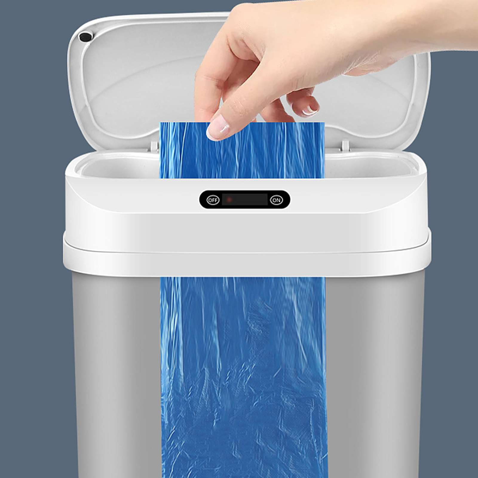 Eine Hand legt einen blauen Müllbeutel in einen weißen und silberfarbenen automatischen Mülleimer. Der Deckel ist offen.