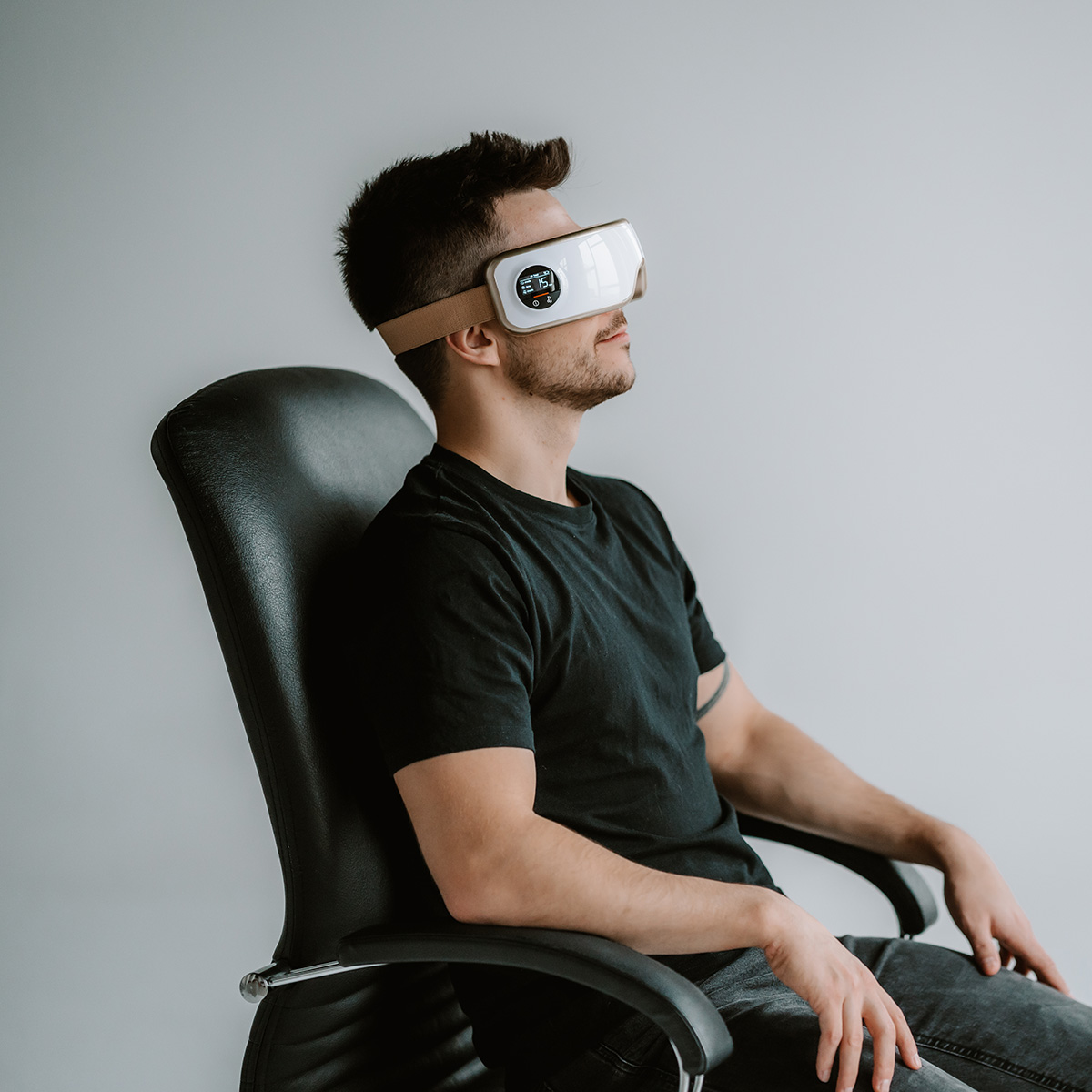 Ein Mann mit Augenmassagegerät sitzt in einem schwarzen Stuhl und hat die Augen geschlossen. Er trägt ein schwarzes T-Shirt und graue Jeans.