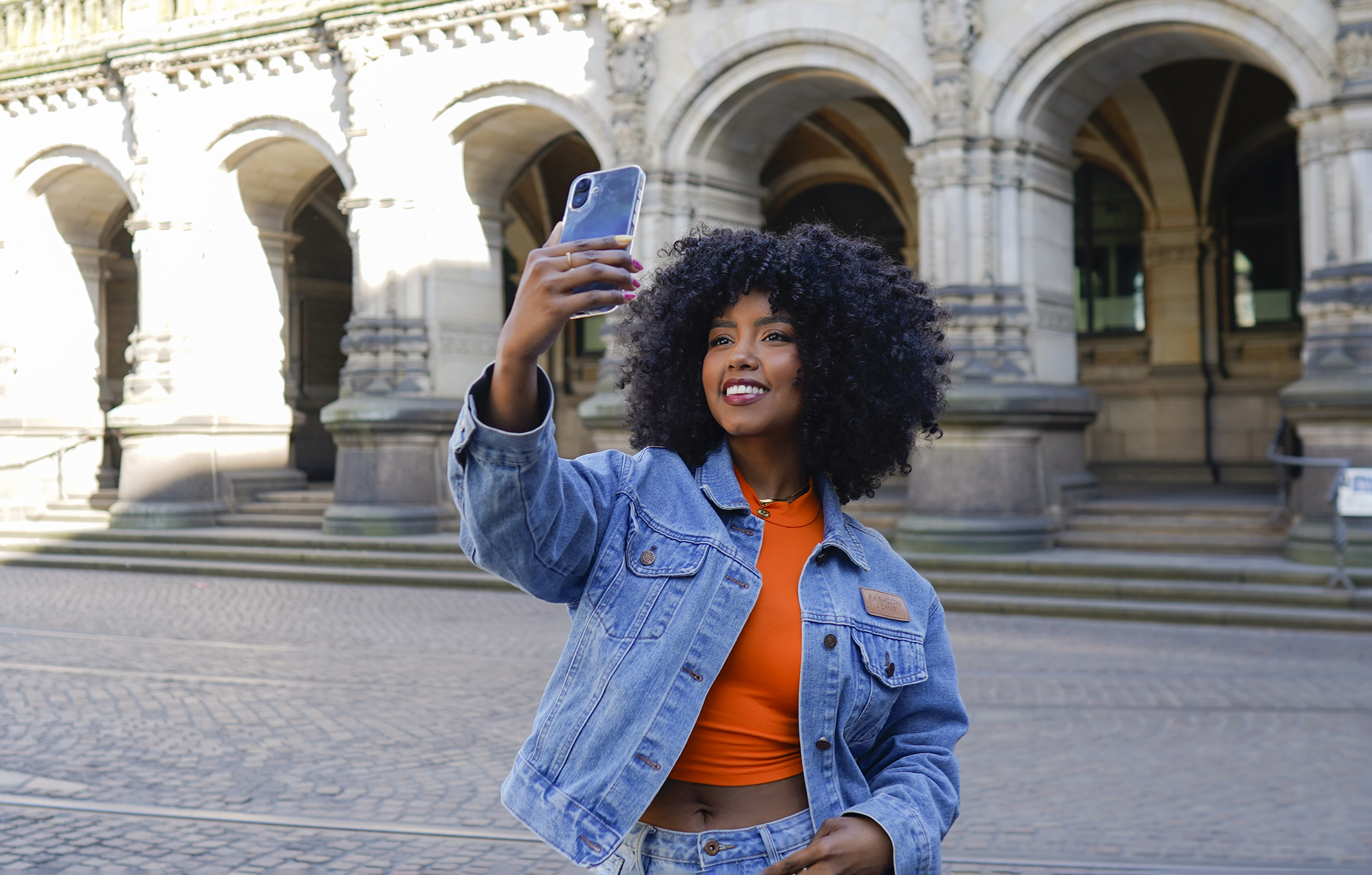 Eine Frau macht ein Selfie mit einem Telefon. Sie trägt eine Jeansjacke und steht vor einem Gebäude mit Bögen.