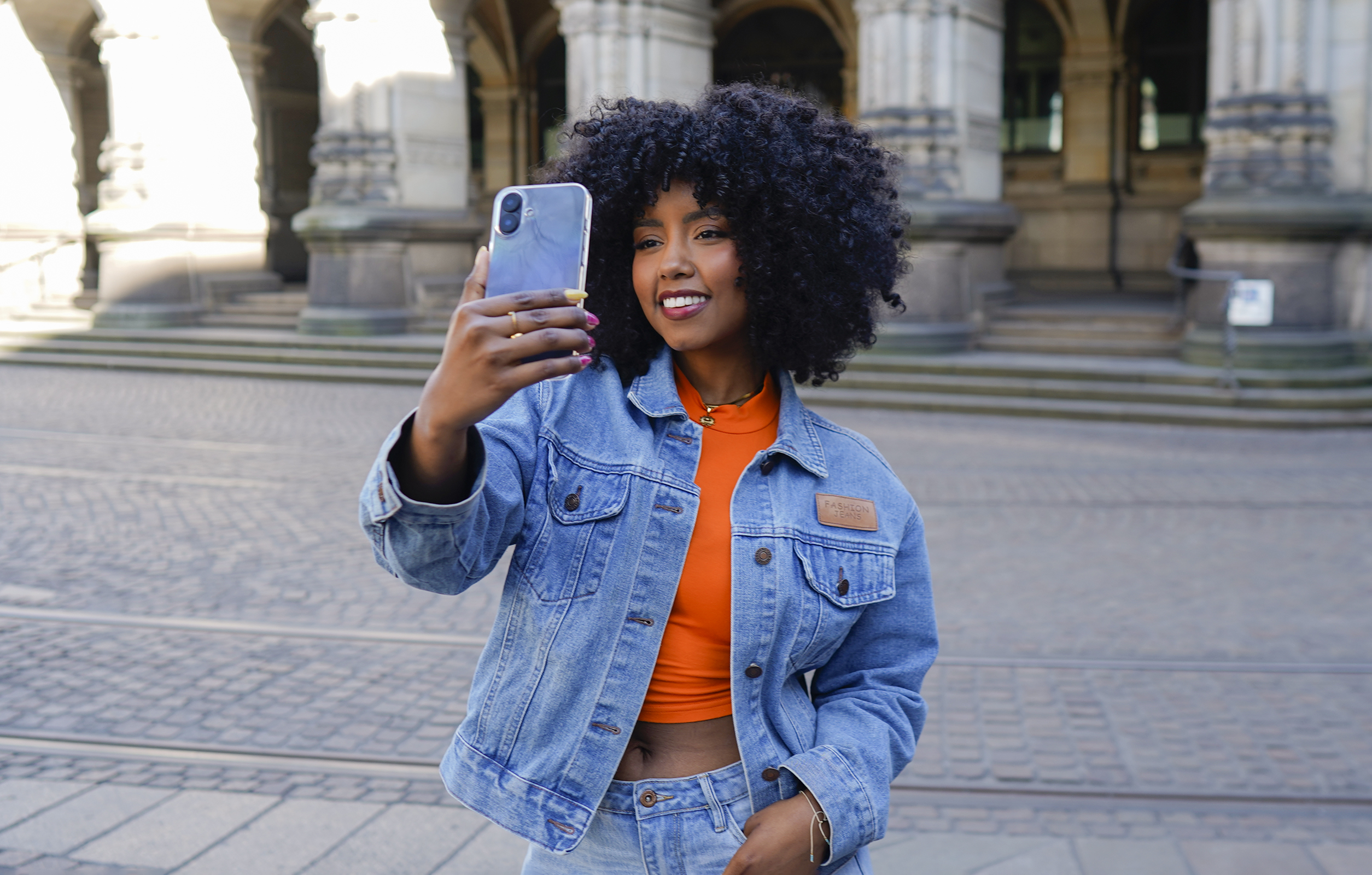 Frau in Jeansjacke macht Selfie mit Handy. Sie lächelt. Im Hintergrund ein Gebäude.