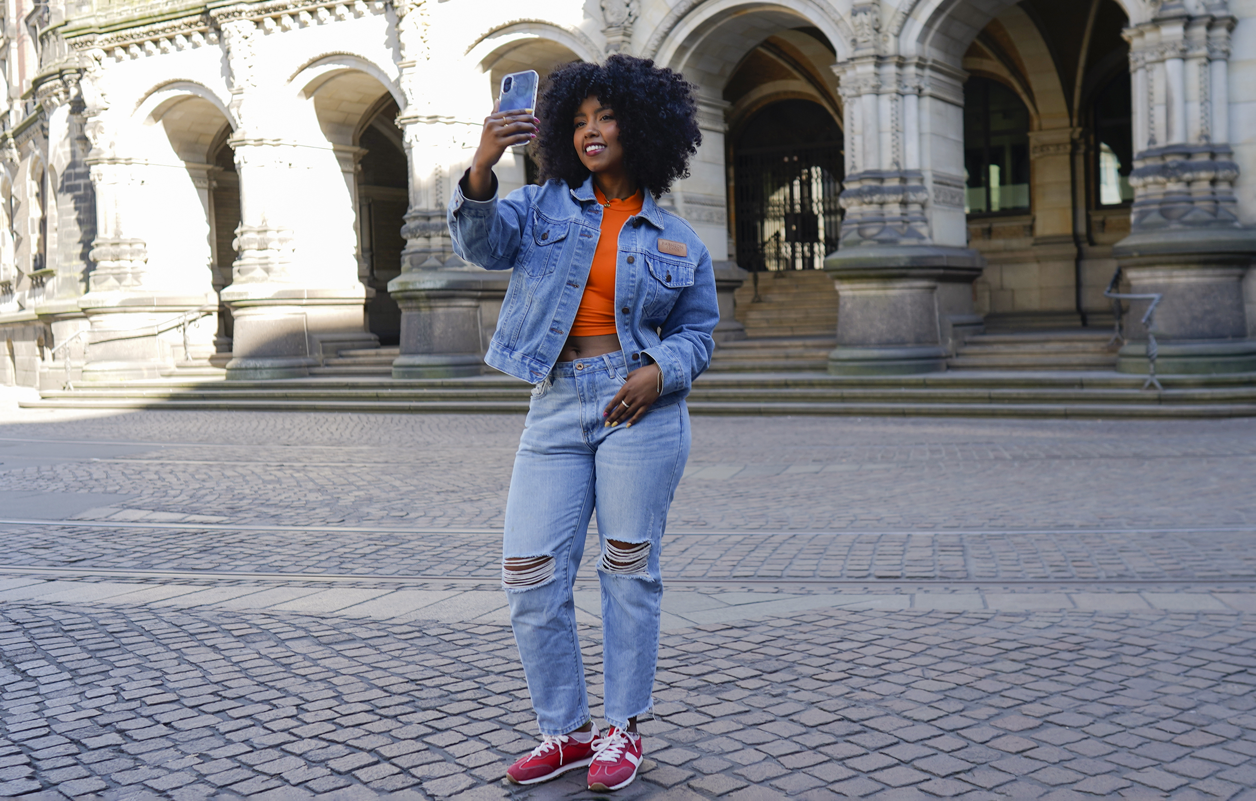 Frau in Jeansjacke und zerrissenen Jeans macht Selfie vor einem Gebäude mit Bögen.