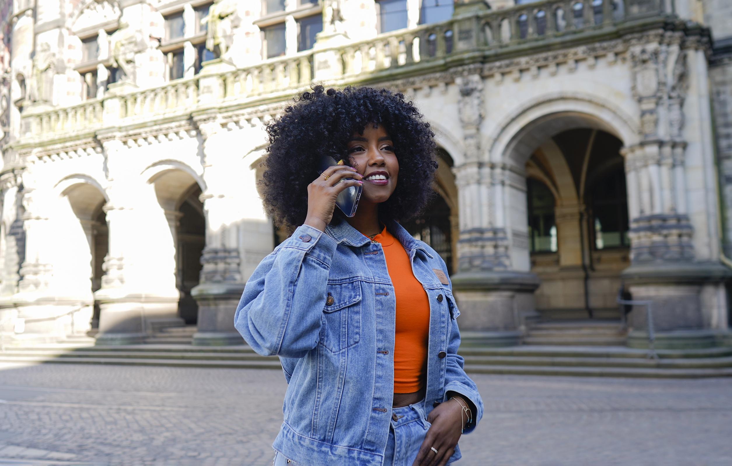 Frau in Jeansjacke telefoniert. Gebäude mit Bögen im Hintergrund.