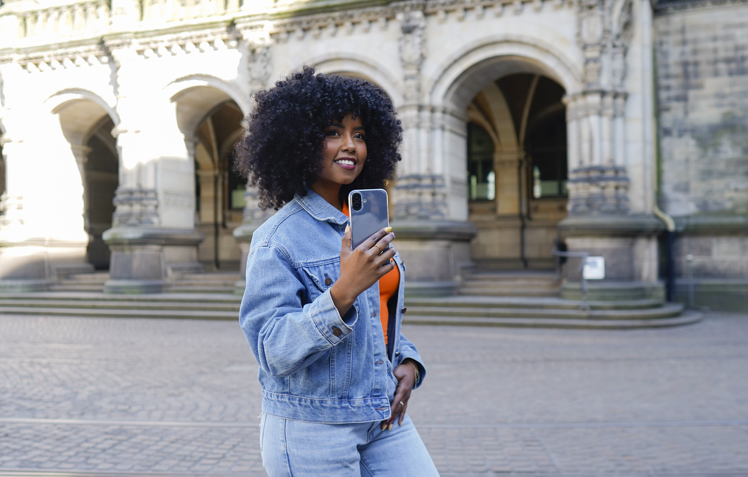 Frau in Jeansjacke, die ein Telefon hält, lächelt. Gebäude mit Bögen im Hintergrund.