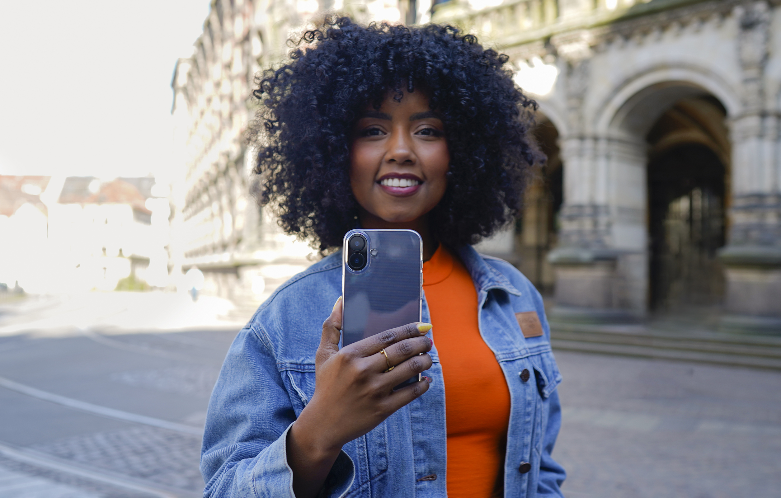 Eine Frau mit lockigem Haar hält ein Telefon. Sie trägt ein orangefarbenes Hemd und eine Jeansjacke und lächelt. Im Hintergrund ist ein Gebäude.