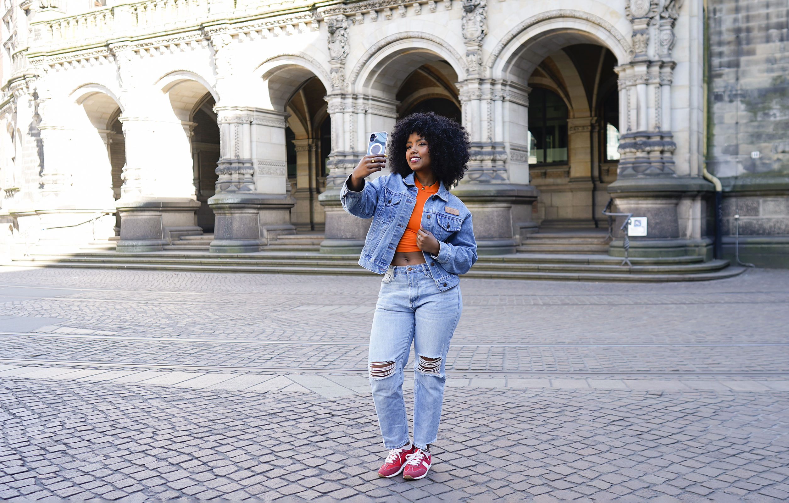 Frau in Jeansjacke und Jeans macht Selfie vor einem Gebäude mit Bögen.
