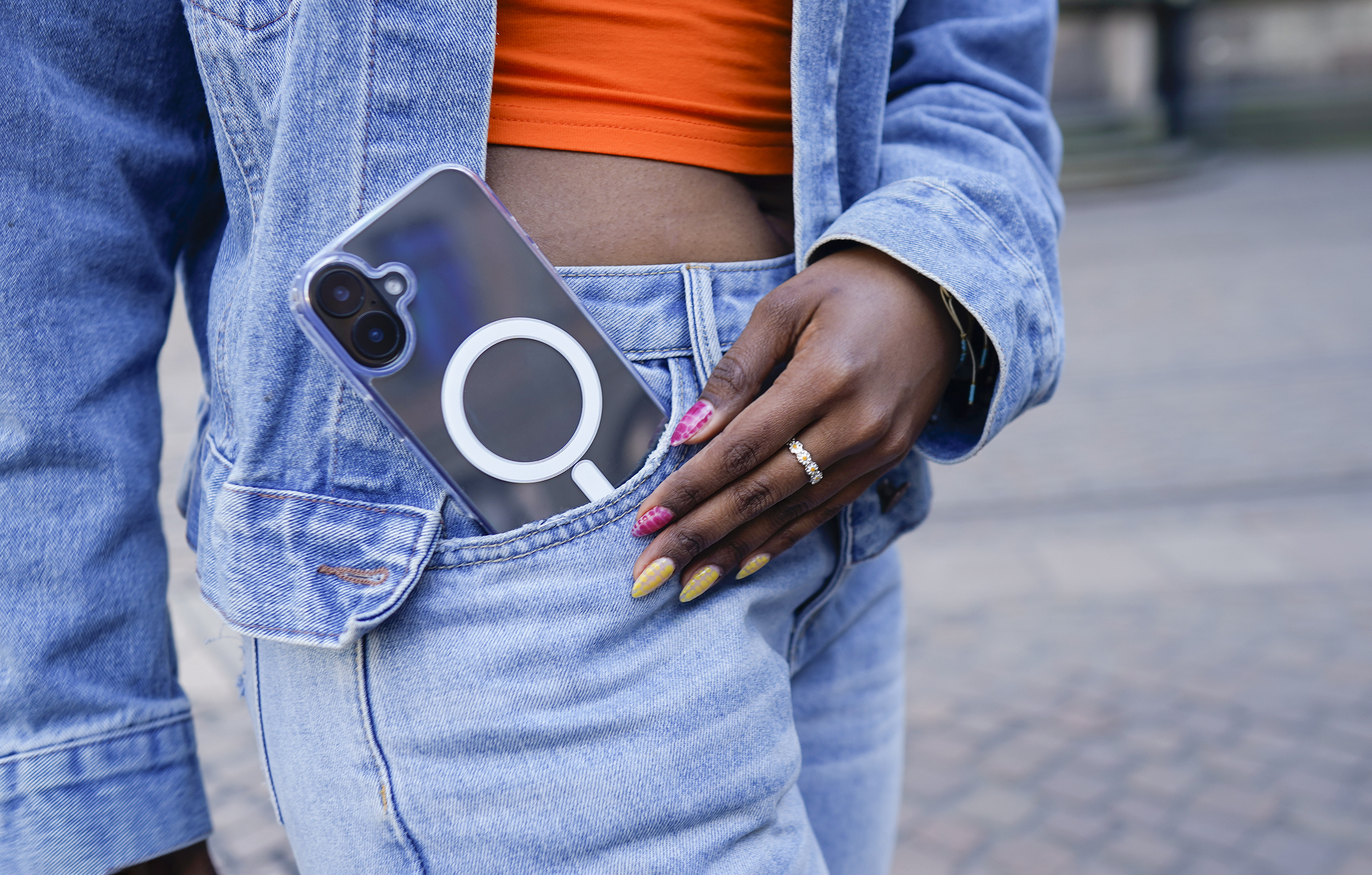 Person in Jeansjacke und -hose, hält Telefon in der Tasche. Person hat Ring und Nagellack.