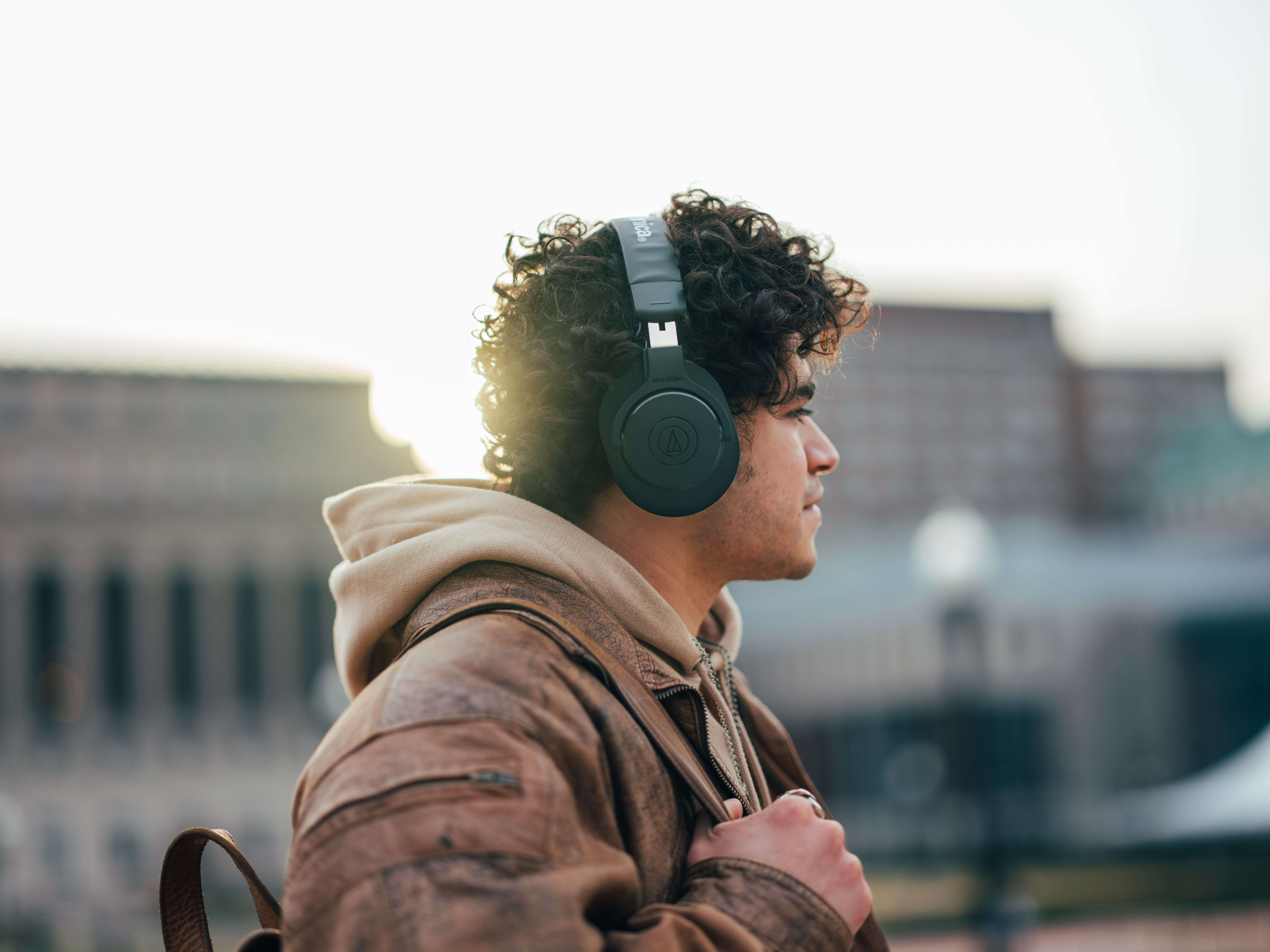 Mężczyzna z kręconymi włosami w słuchawkach i skórzanej kurtce, patrzący w prawo. Budynki w tle.
