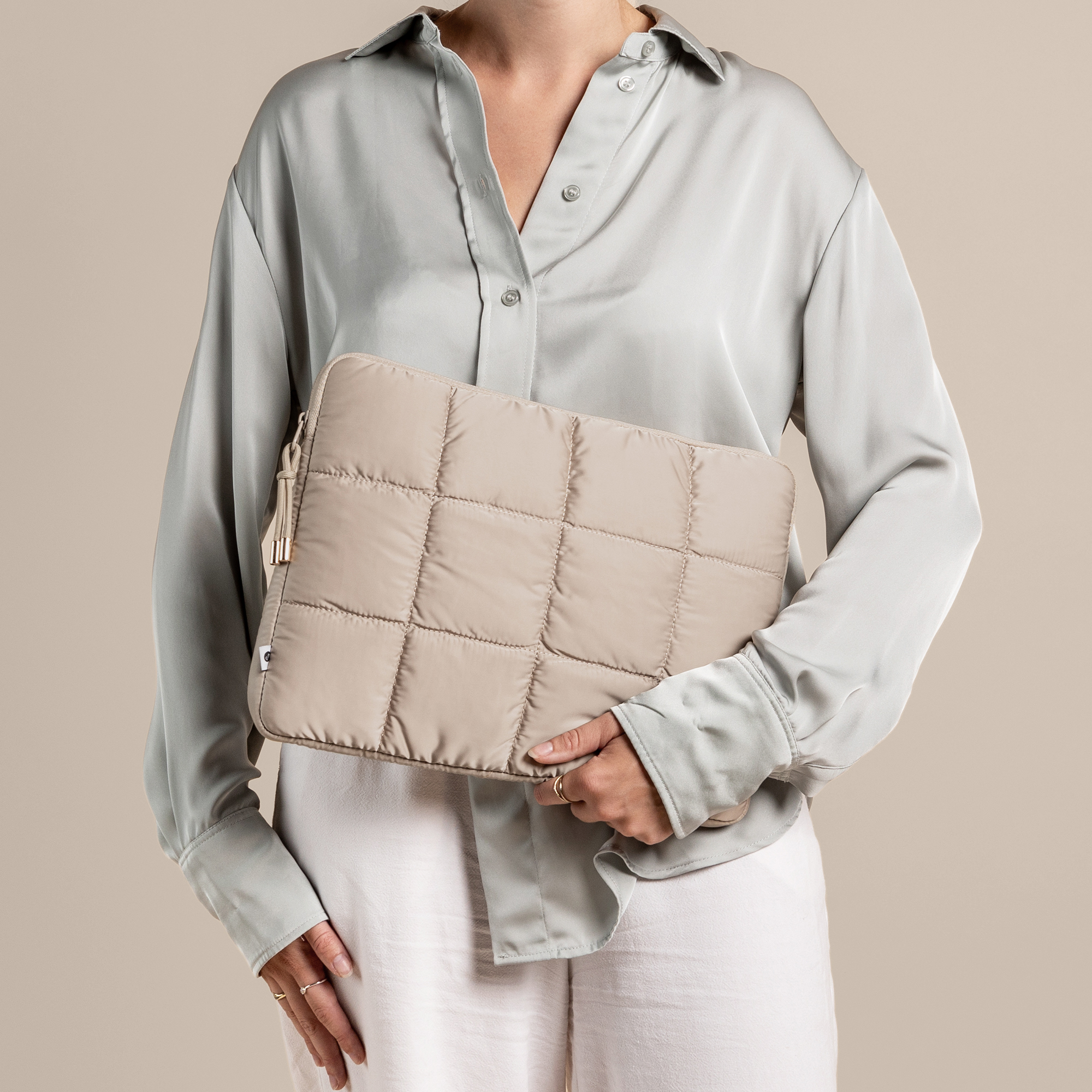 A person holds a beige padded laptop sleeve, dressed in a light green shirt and white pants, neutral background.