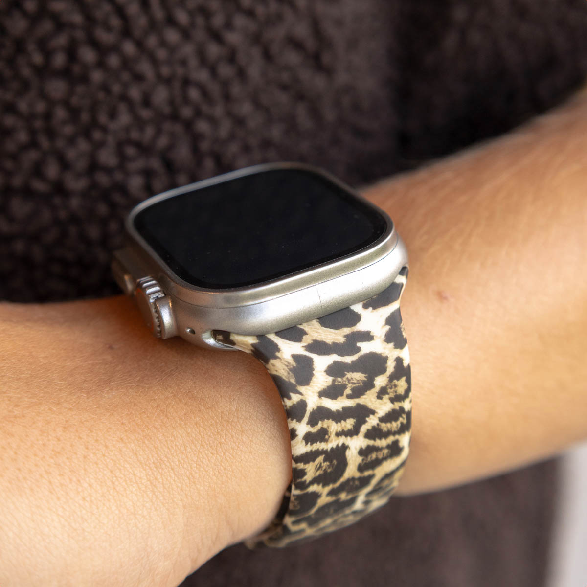 A close-up shows a wrist with a leopard print watch band and a silver watch. The background is a fuzzy brown sweater.