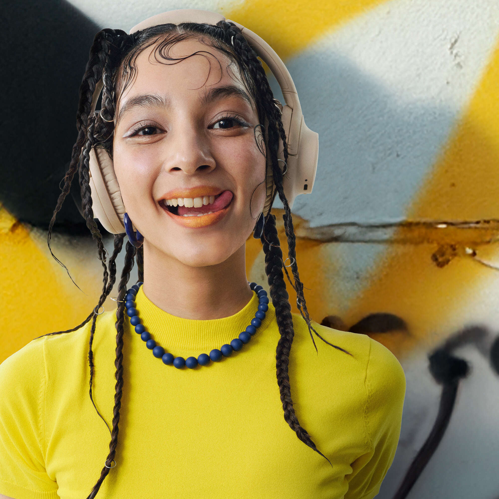 Giovane donna con trecce, cuffie e top giallo. Tira fuori la lingua davanti a uno sfondo di graffiti.