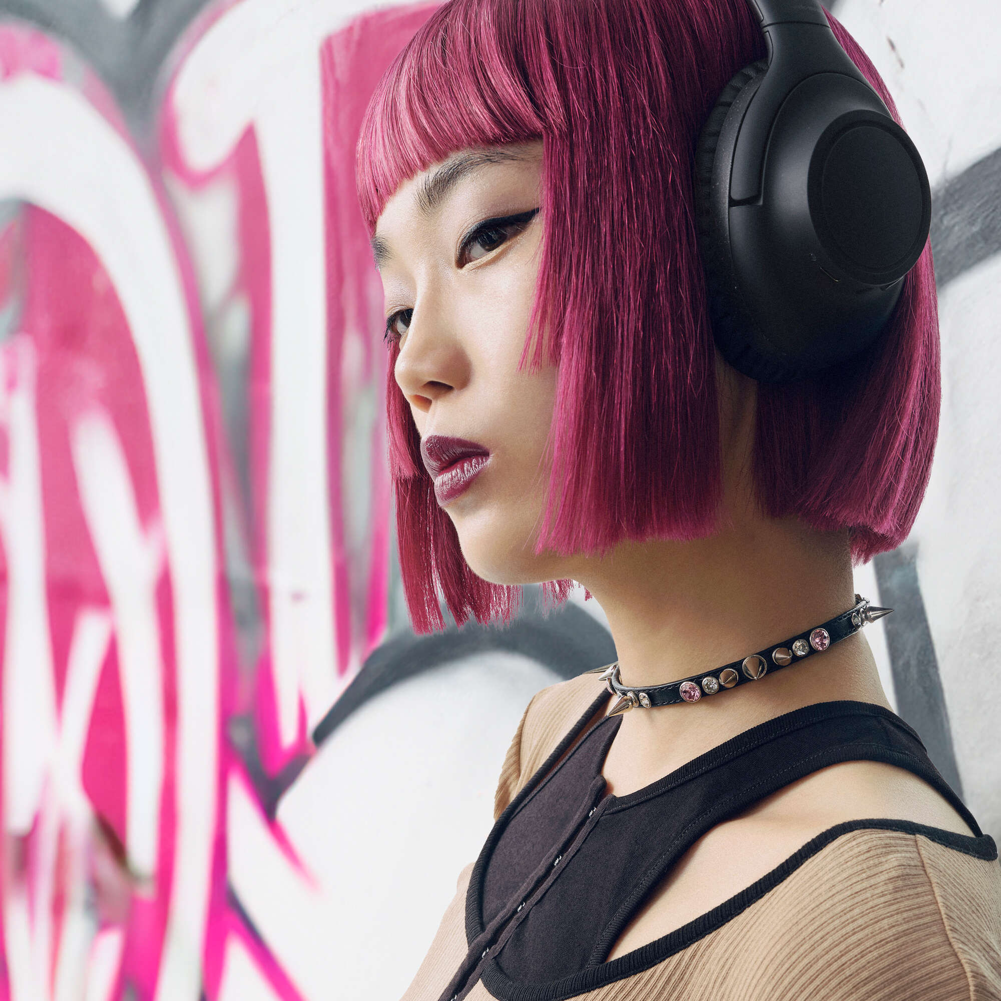Osoba z różowymi włosami i słuchawkami stoi przed graffiti. Ma na sobie kolczastą obrożę i czarny top.