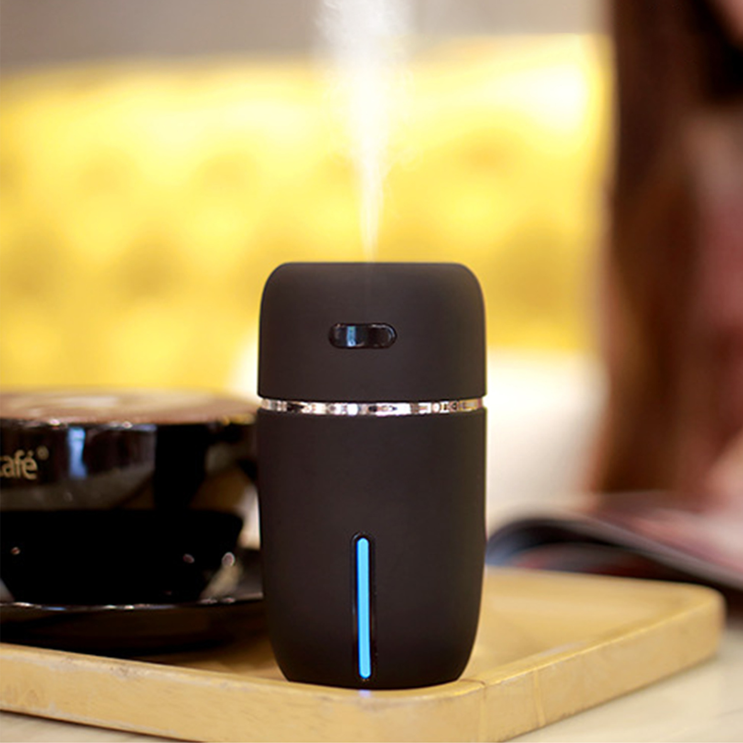 Czarny nawilżacz uwalnia parę. Obok na drewnianej tacy stoi filiżanka kawy i czasopismo. Tło jest rozmyte żółte.