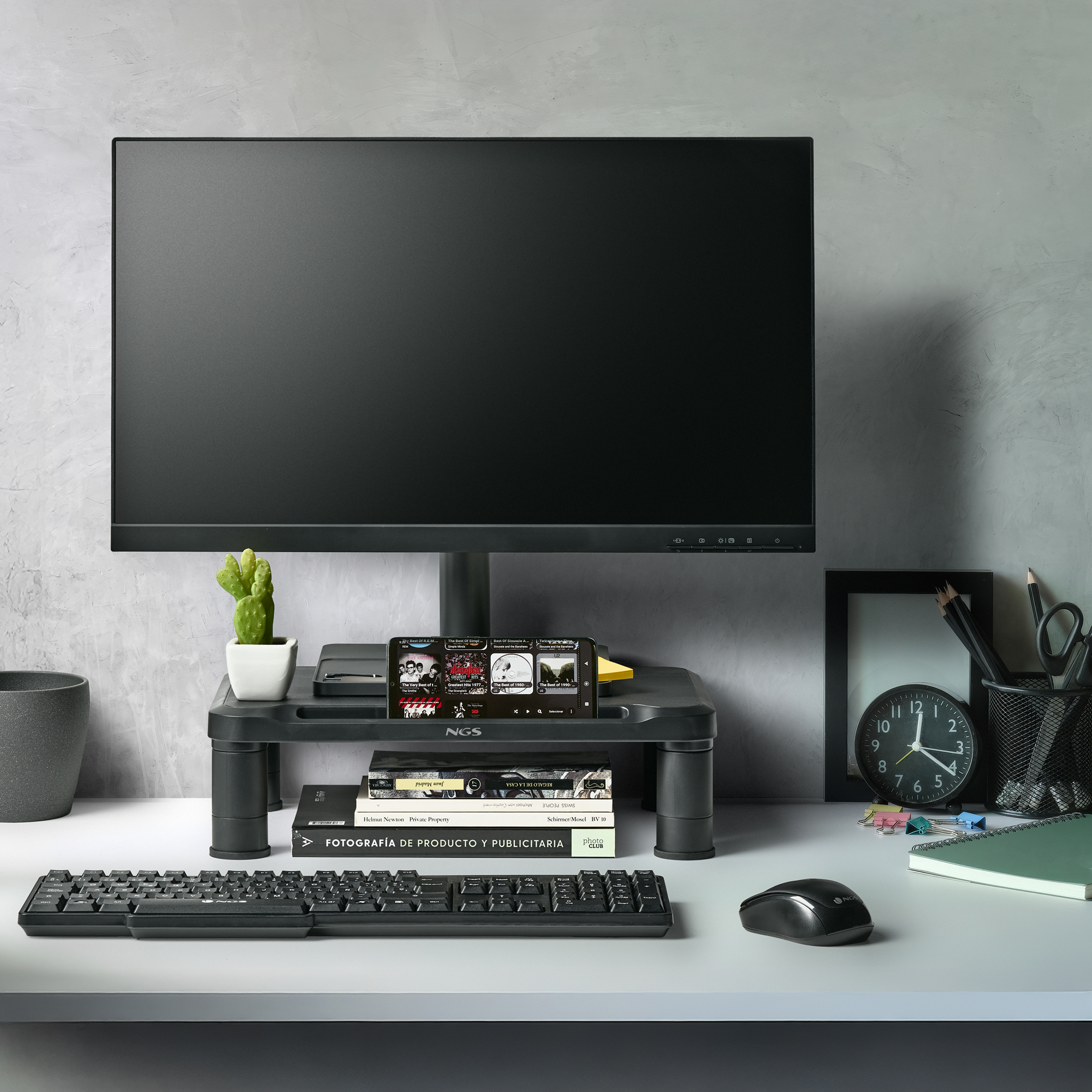 Schreibtisch mit Monitor, Tastatur, Maus, Kaktus, Uhr und Büchern. Ein Telefon zeigt Musik.