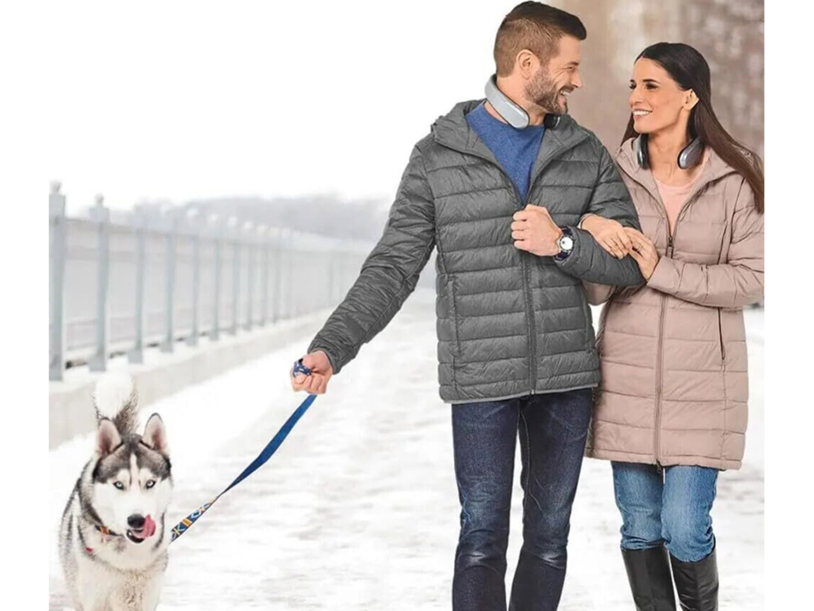 Una pareja pasea con un husky por un camino nevado. El hombre sostiene la correa. Llevan chaquetas.