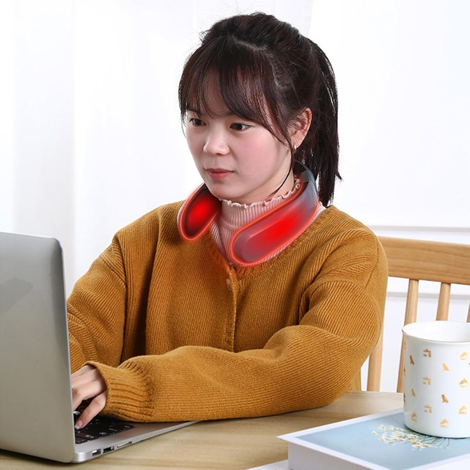 Una mujer con un masajeador de cuello está sentada en un escritorio con una laptop y una taza. Lleva un suéter amarillo.