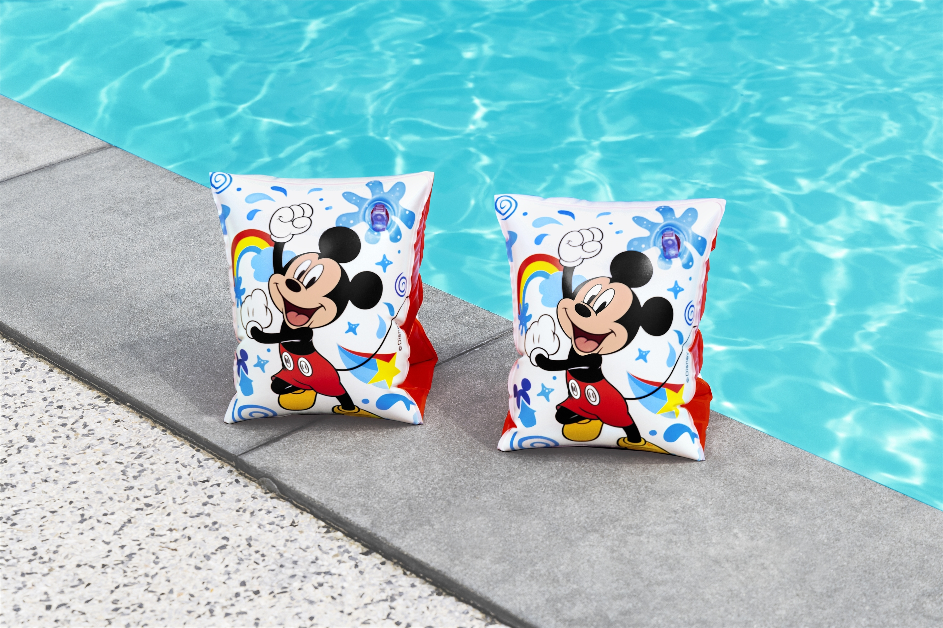 Zwei Mickey-Mouse-Arm-Schwimmflügel liegen am Pool. Der Hintergrund ist blaues Wasser. Die Schwimmflügel haben Regenbogen und Sterne.