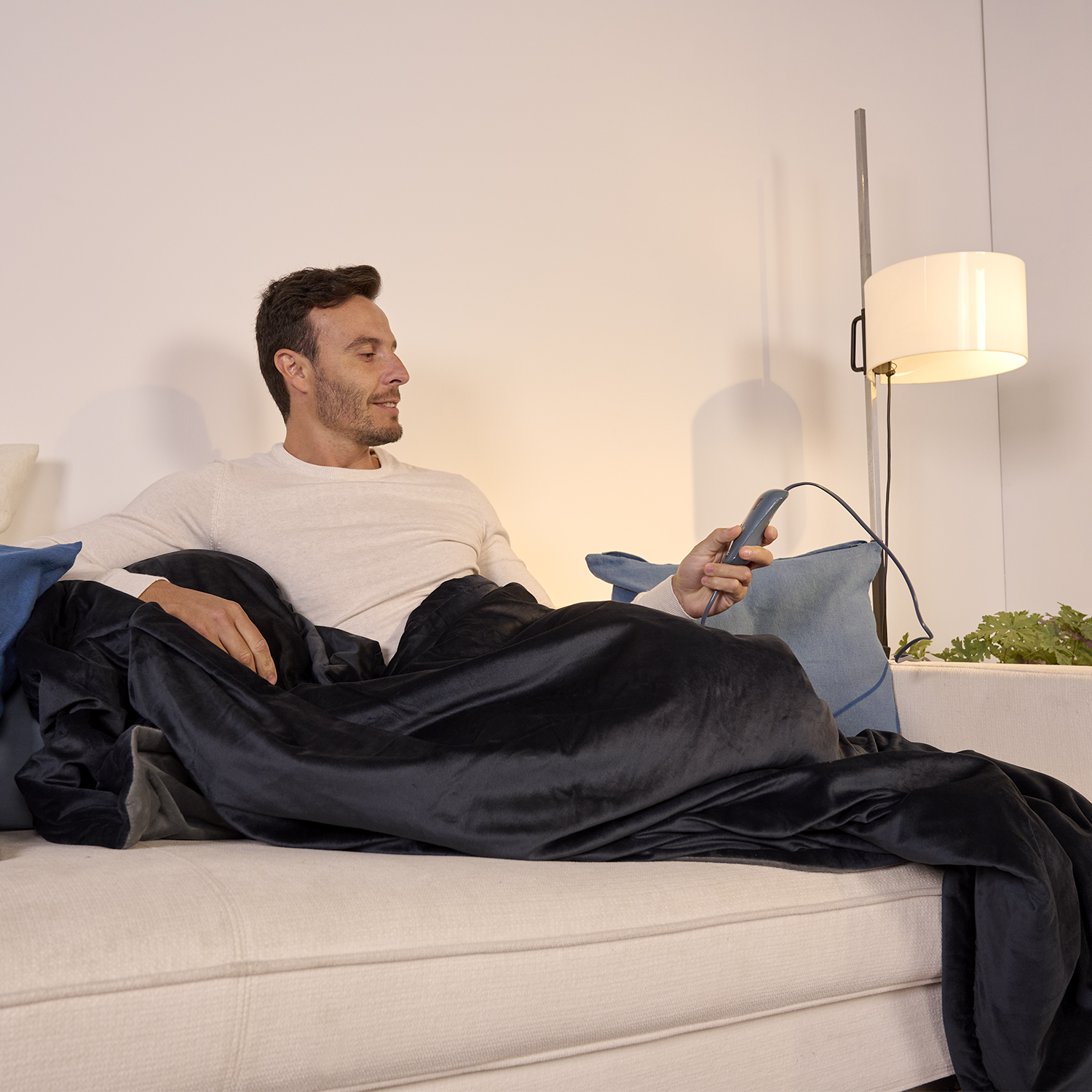 Mężczyzna siedzi na kanapie, przykryty czarnym kocem. Trzyma pilota. Lampa jest włączona. Tło to biała ściana.