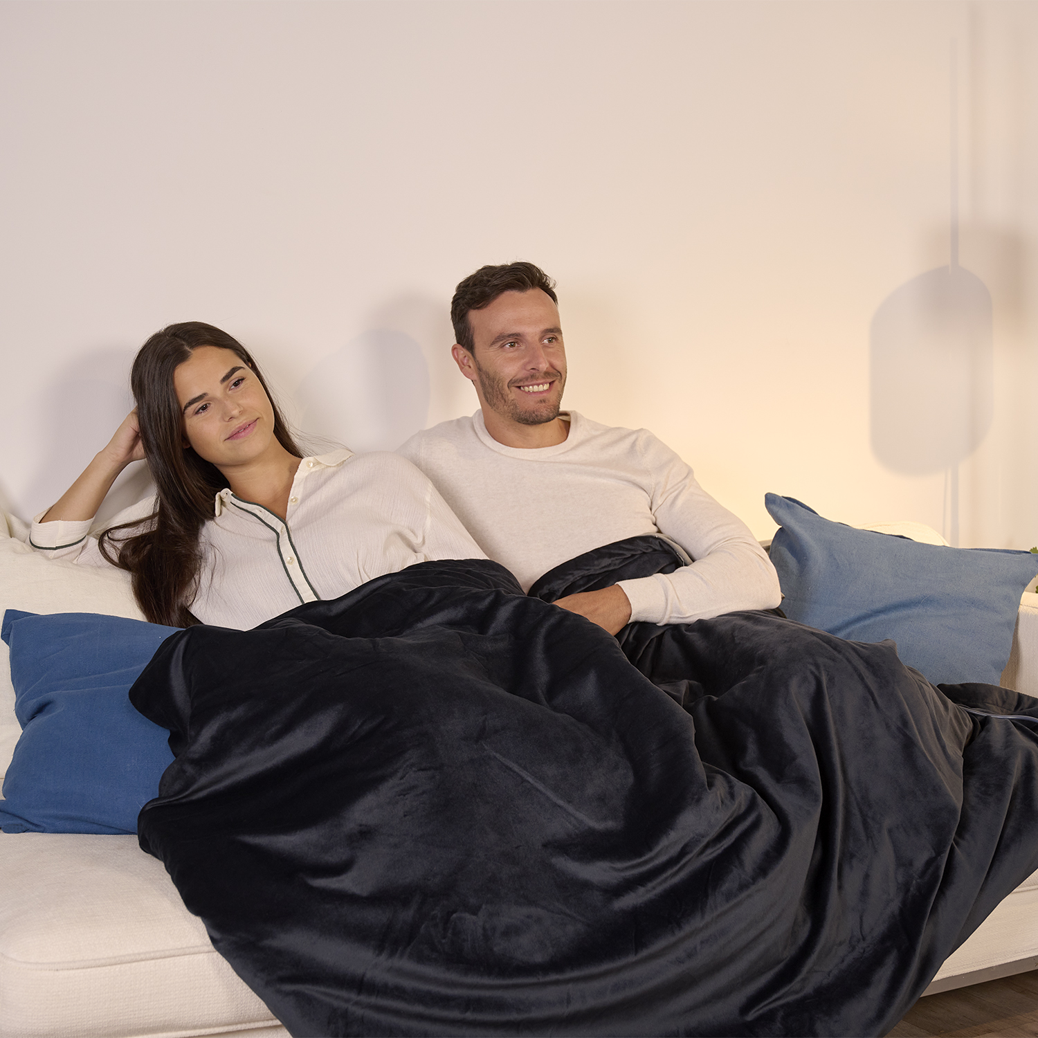 Para siedzi na białej kanapie, przykryta czarnym kocem. Kobieta opiera głowę na dłoni, mężczyzna się uśmiecha. Widoczne są niebieskie poduszki.
