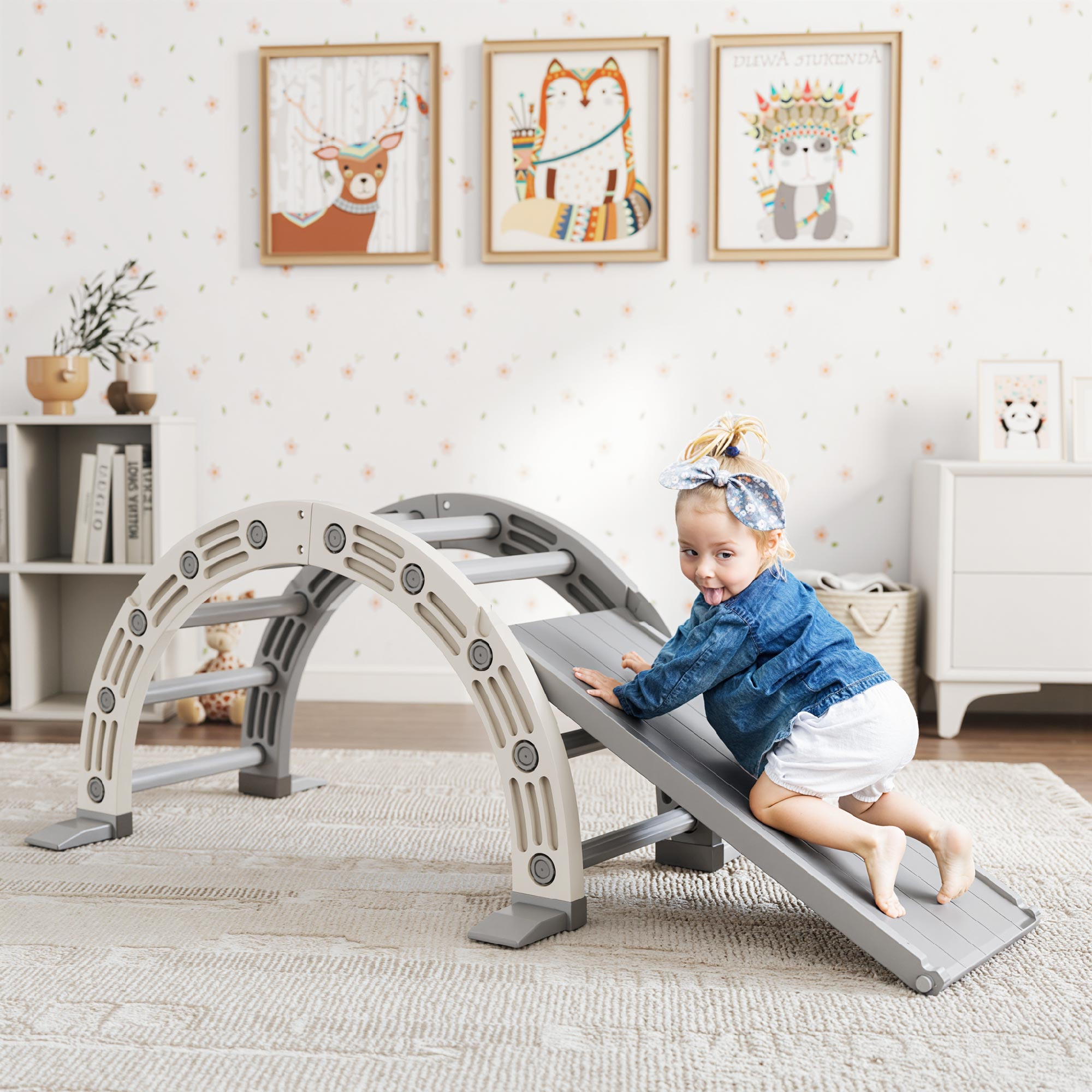 Un niño pequeño con chaqueta vaquera y pantalones cortos blancos escala una estructura de juego gris con un tobogán en una habitación con arte.