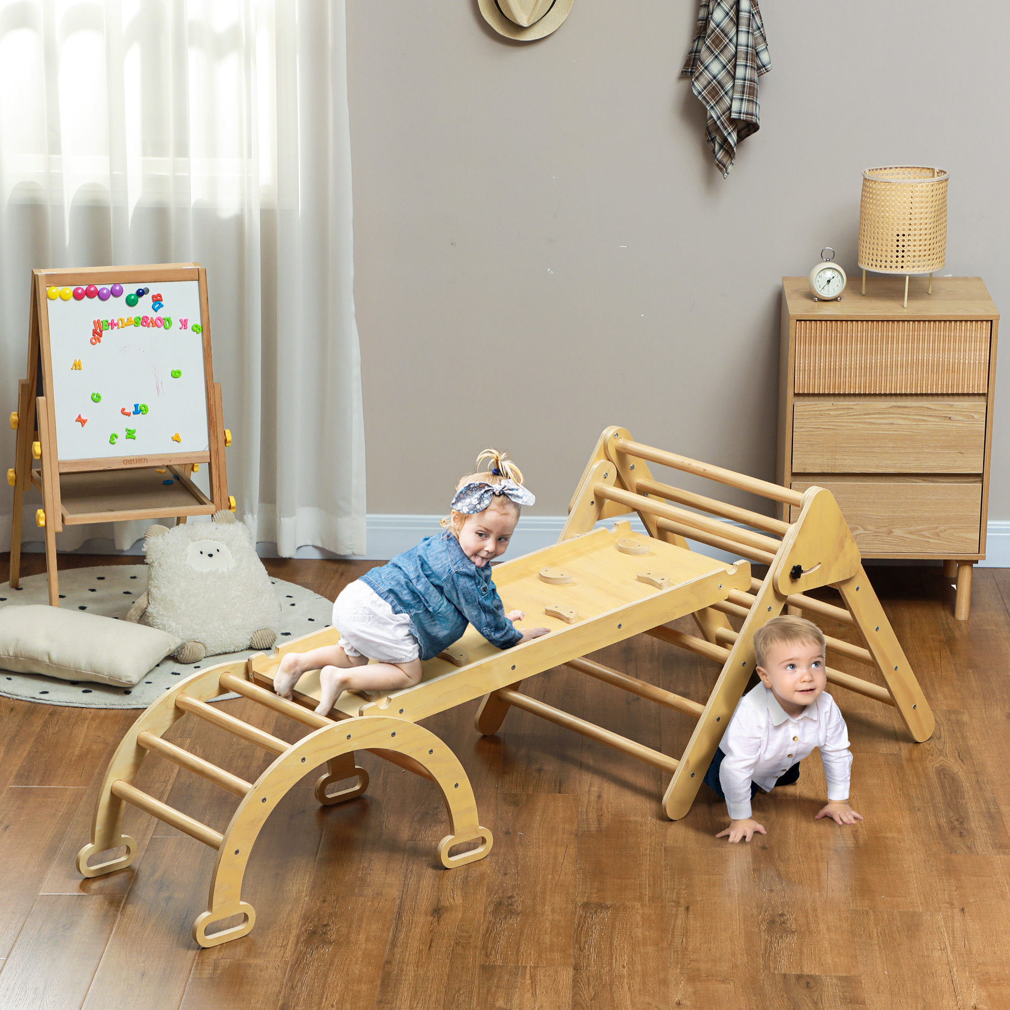 Dos niños pequeños juegan en una estructura de escalada de madera en interiores. Uno escala, el otro gatea. Una pizarra y un armario están en el fondo.