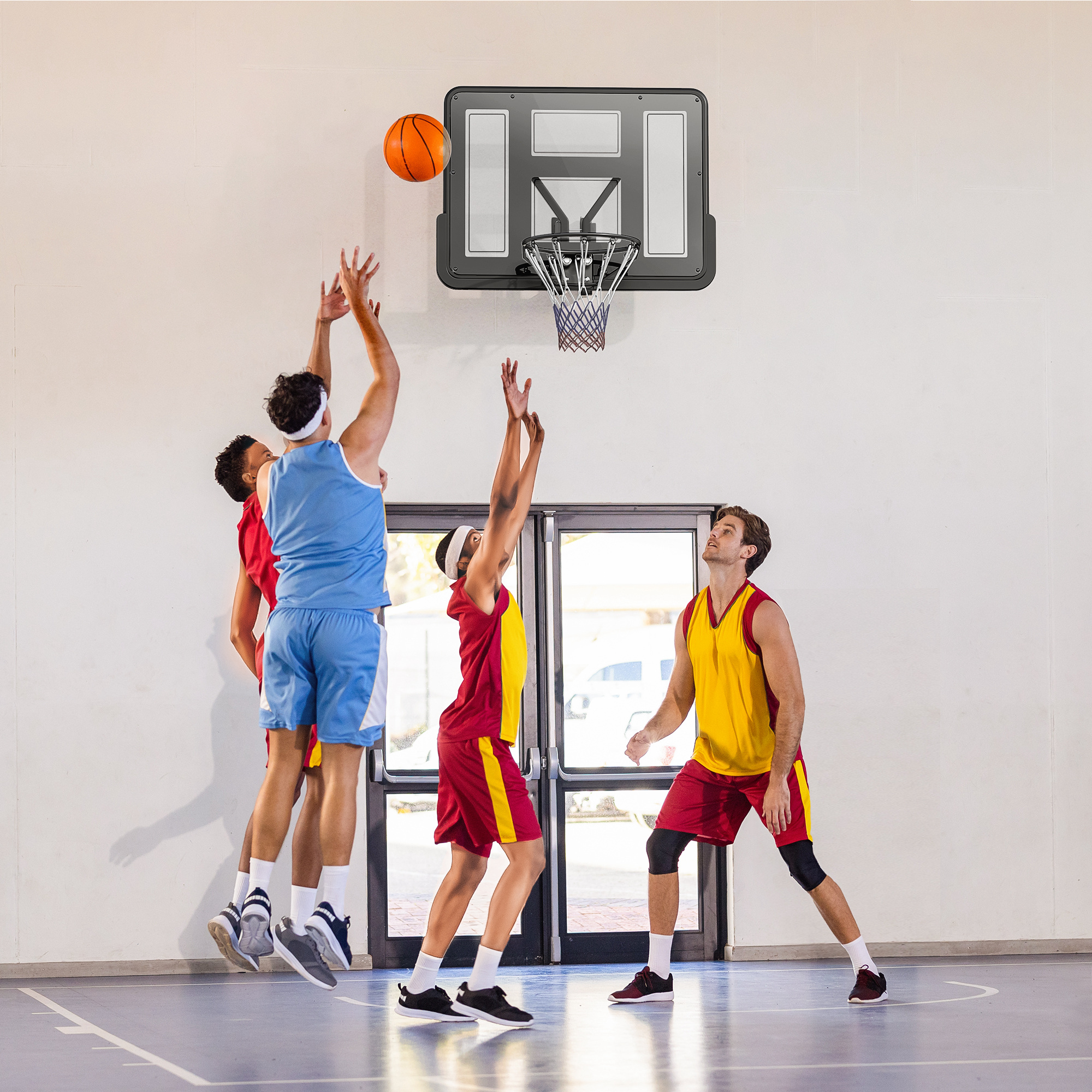 Partido de baloncesto en curso. Jugadores en acción, saltando por el balón, apuntando al aro. Cancha con pared blanca al fondo.