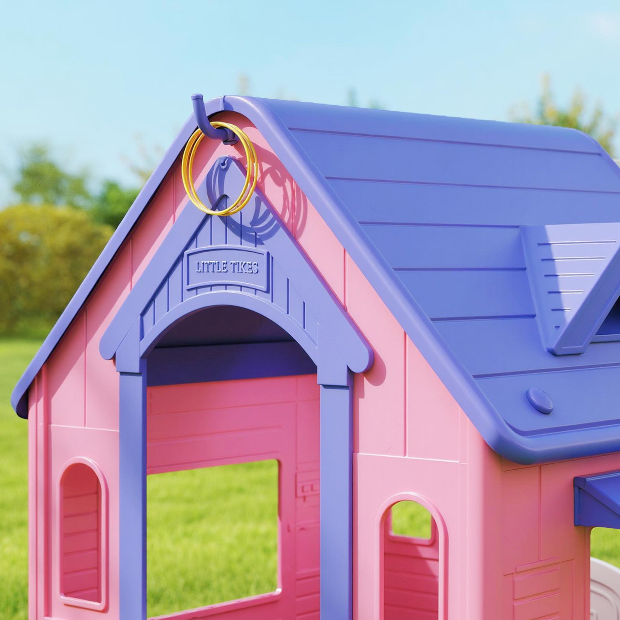 Una casita de juegos Little Tikes rosa y morada. Un anillo amarillo cuelga del techo. Hierba verde y cielo azul al fondo.