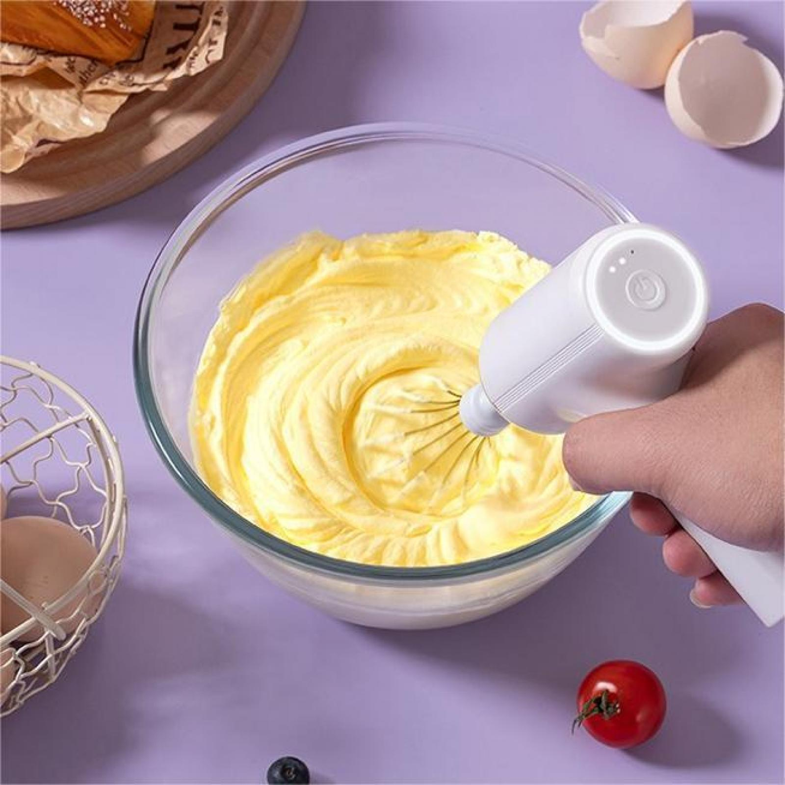 Eine Hand, die einen weißen Mixer hält, vermischt gelben Teig in einer Glasschüssel. Eier und eine Tomate sind in der Nähe.