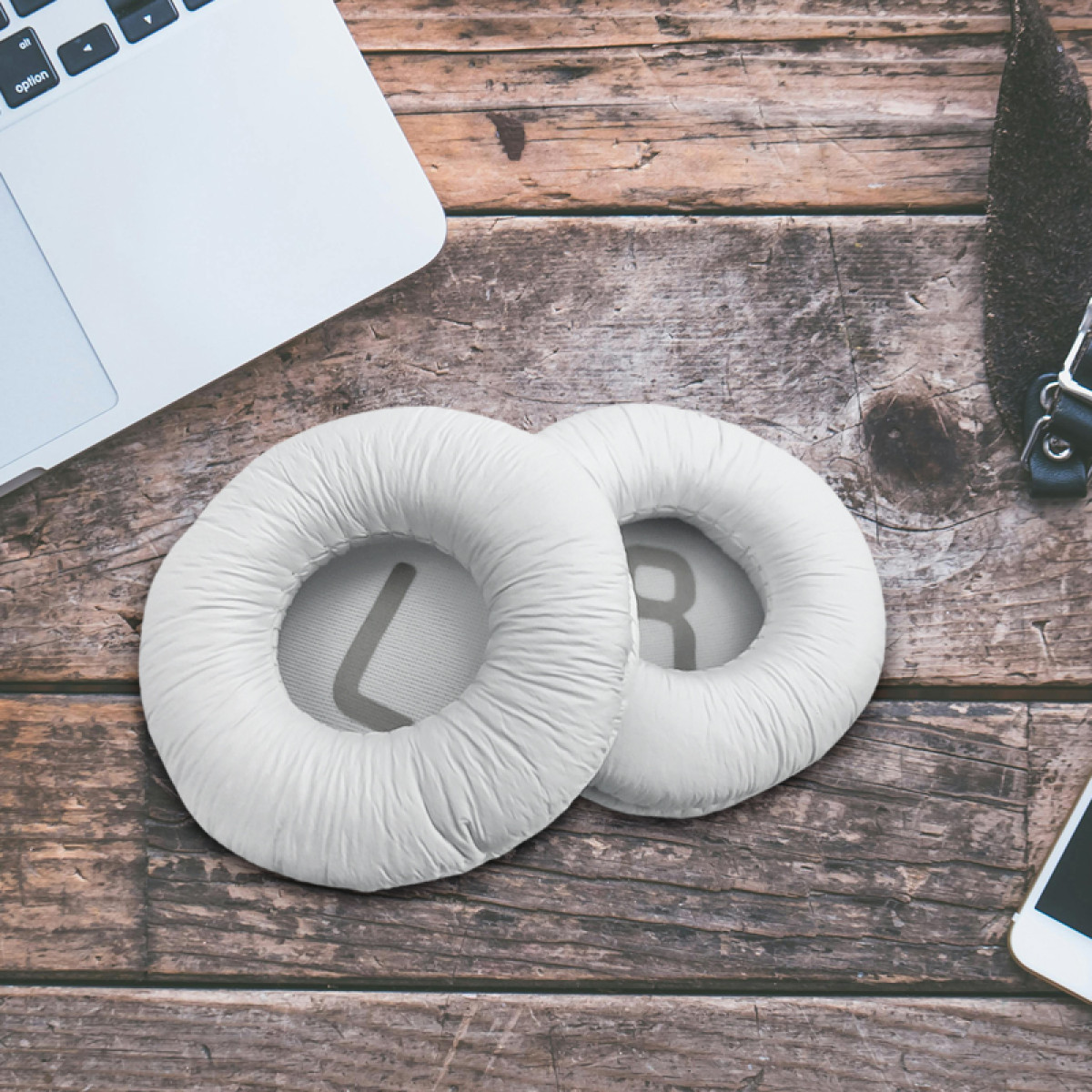 Dwie białe poduszki nauszne do słuchawek na drewnianej powierzchni. W kadrze widoczny jest również laptop i telefon.