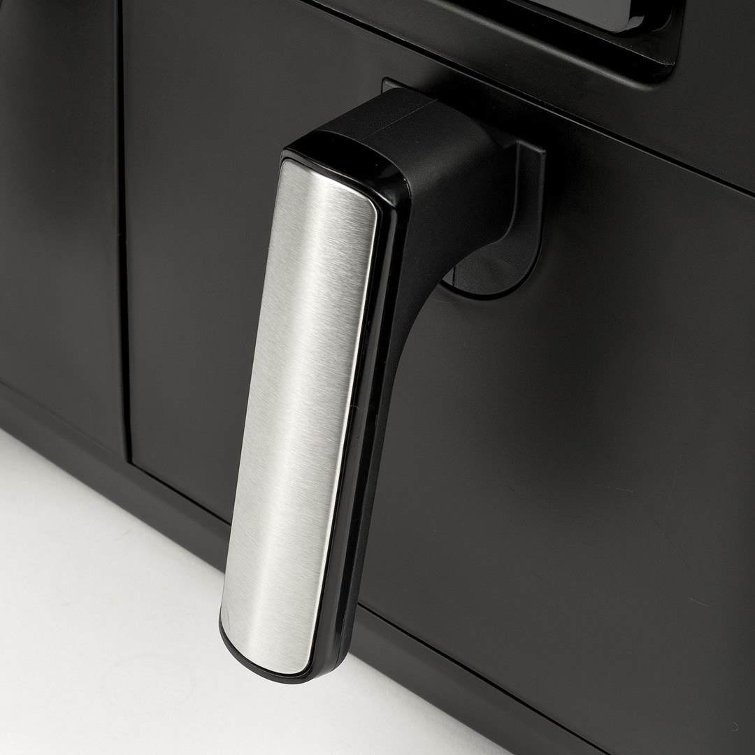 Zbliżenie czarnej frytkownicy powietrznej ze srebrnym uchwytem. Uchwyt znajduje się po prawej stronie. Biała powierzchnia.