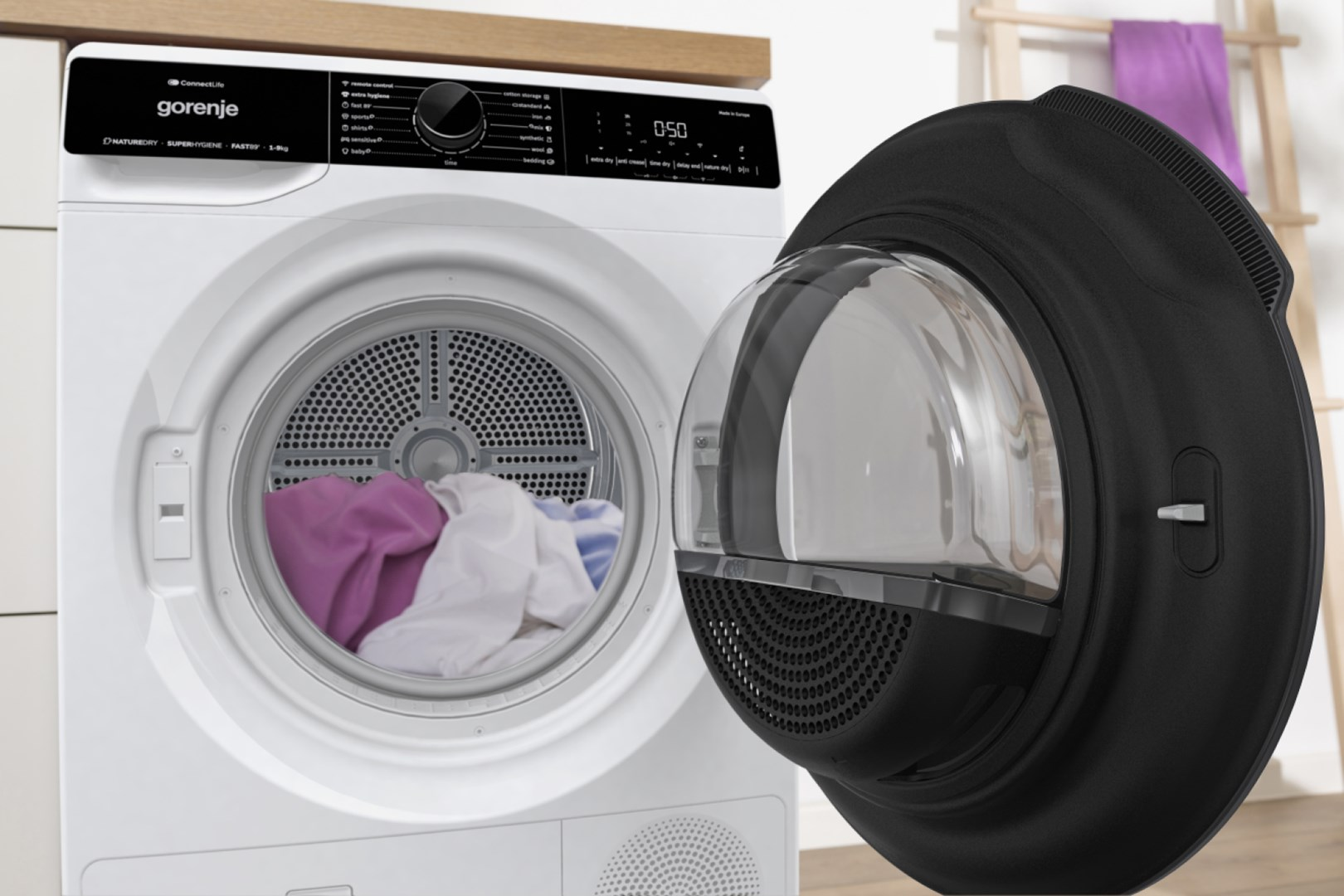 Biała suszarka Gorenje z otwartymi drzwiami, pokazująca ubrania w środku. Czarna rama drzwi. Drewniana drabina z fioletowym ręcznikiem w tle.