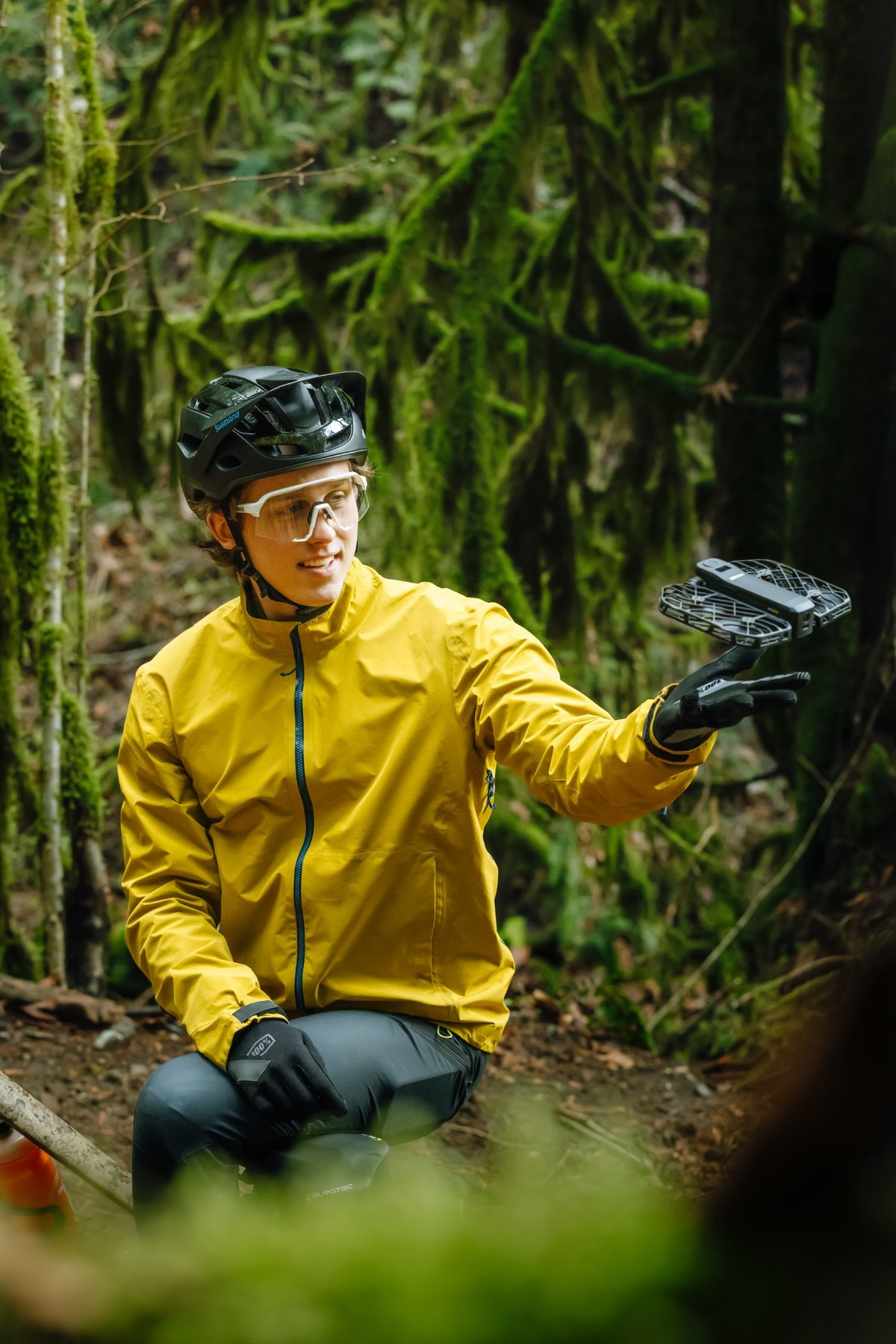 Mężczyzna w żółtej kurtce trzyma drona w lesie. Ma na sobie kask, okulary i rękawice. Zielony mech pokrywa drzewa.