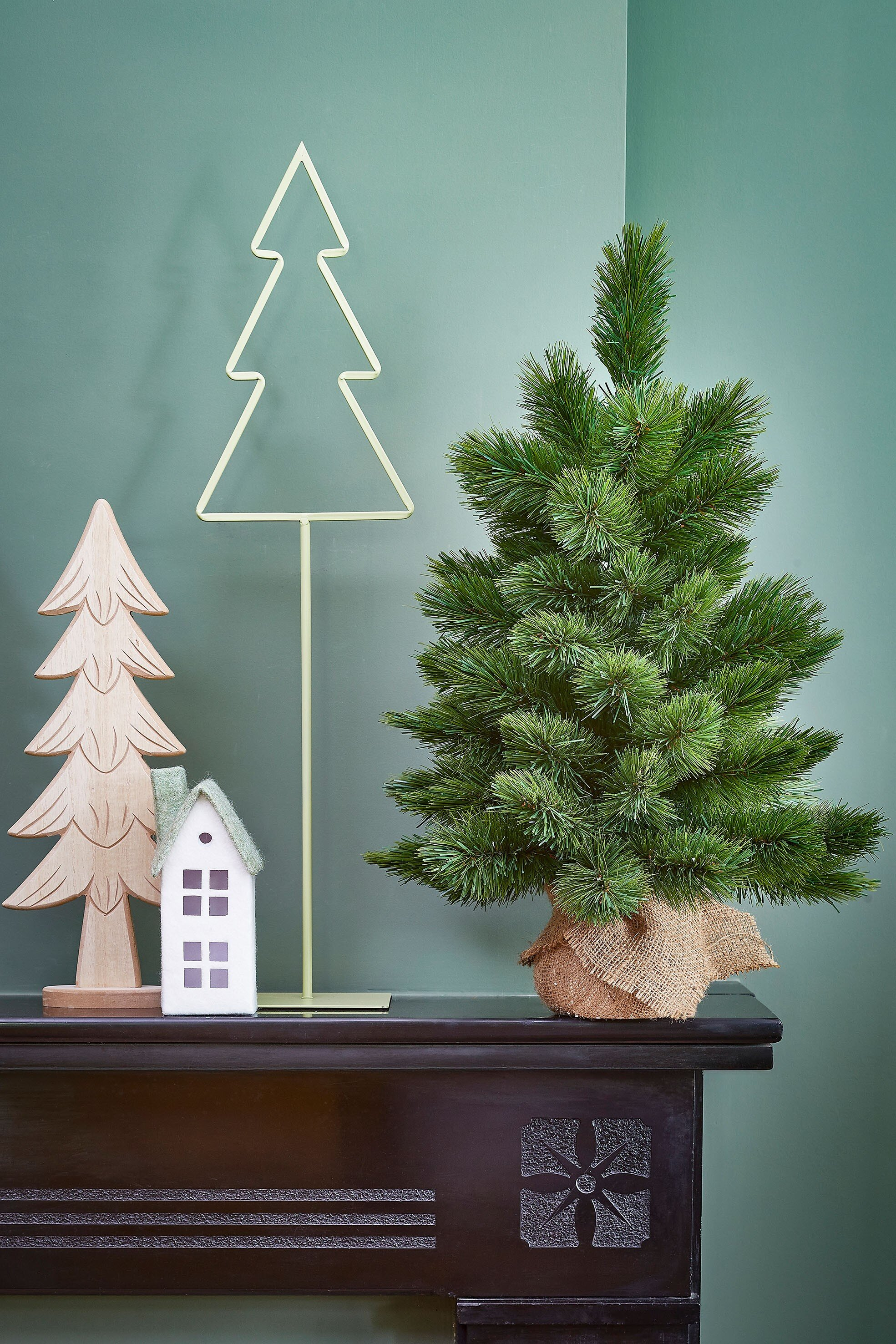Un arbre de Noël vert, un arbre en bois et une maison sur une surface marron foncé contre un mur vert.