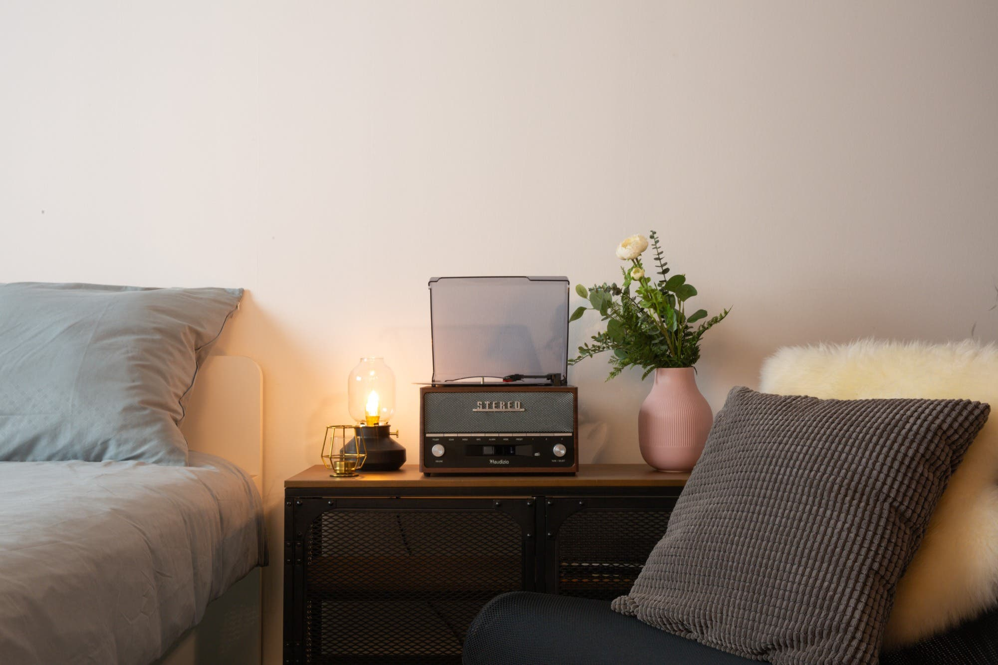 Una escena de dormitorio con un tocadiscos, lámpara, flores y una cama. La paleta de colores es neutra y el ambiente acogedor.