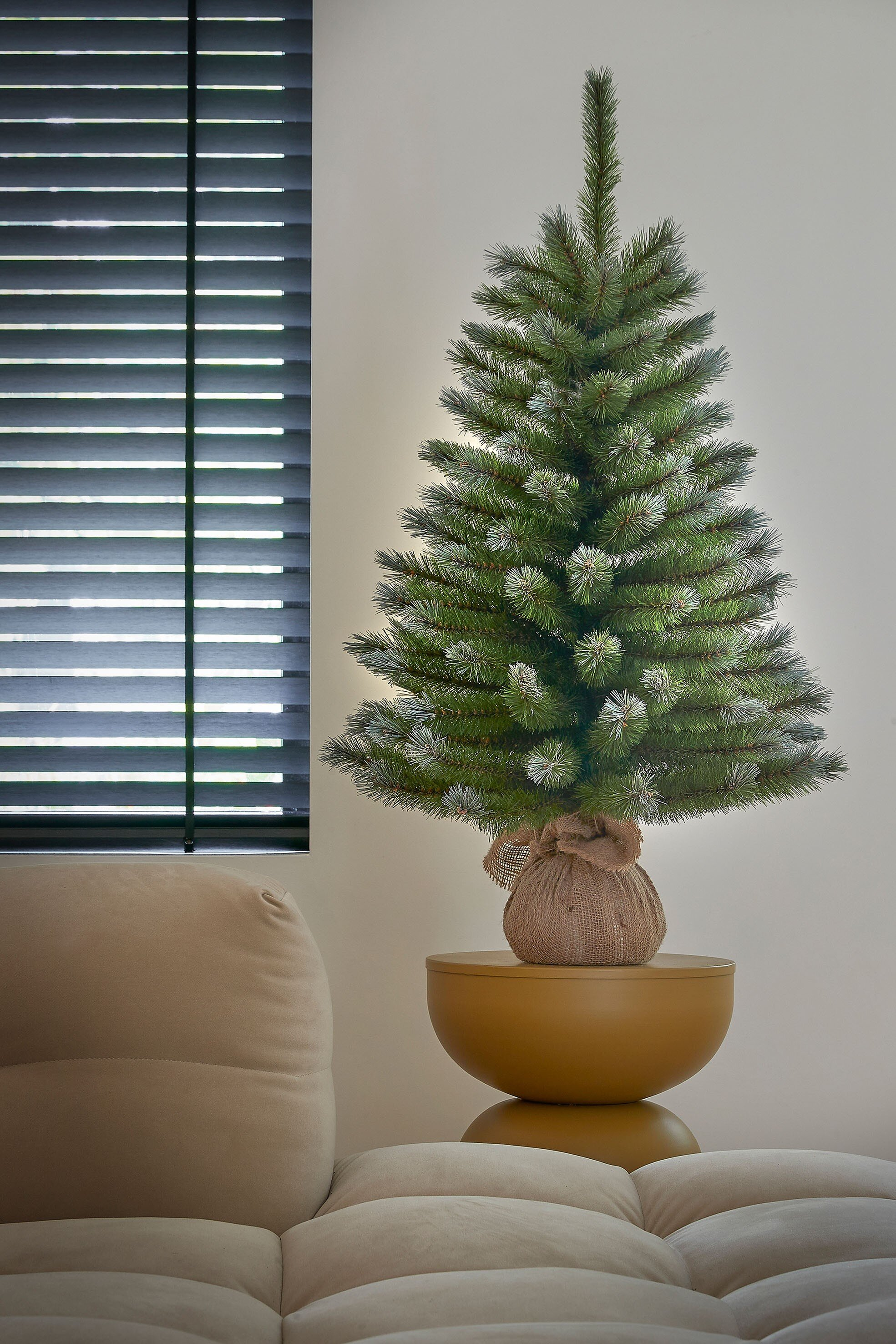 Un petit arbre de Noël sur une table dorée, à côté d'une fenêtre à stores et d'un canapé.