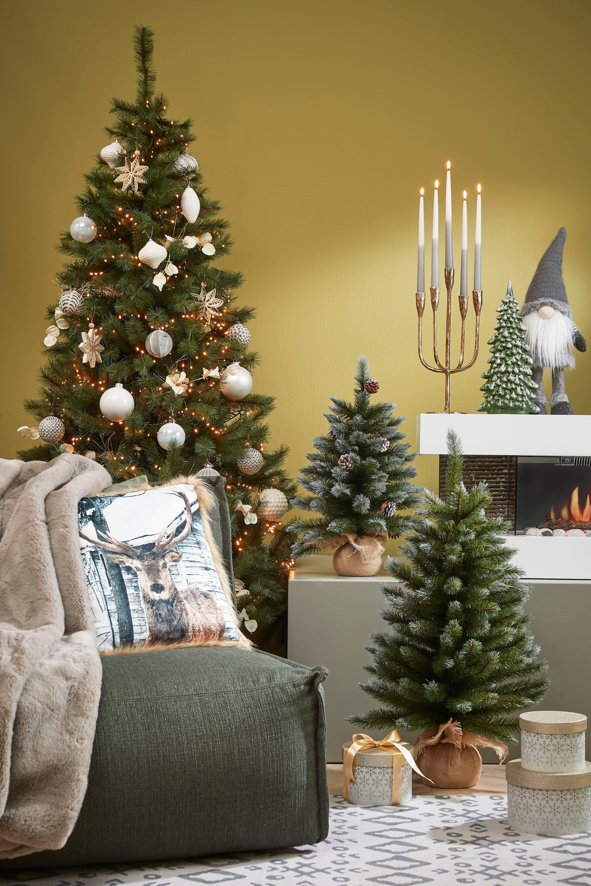 Een versierde kerstboom staat links. Andere bomen, kaarsen en cadeaus zijn in de scène. Een kussen met een hert ligt op een stoel.